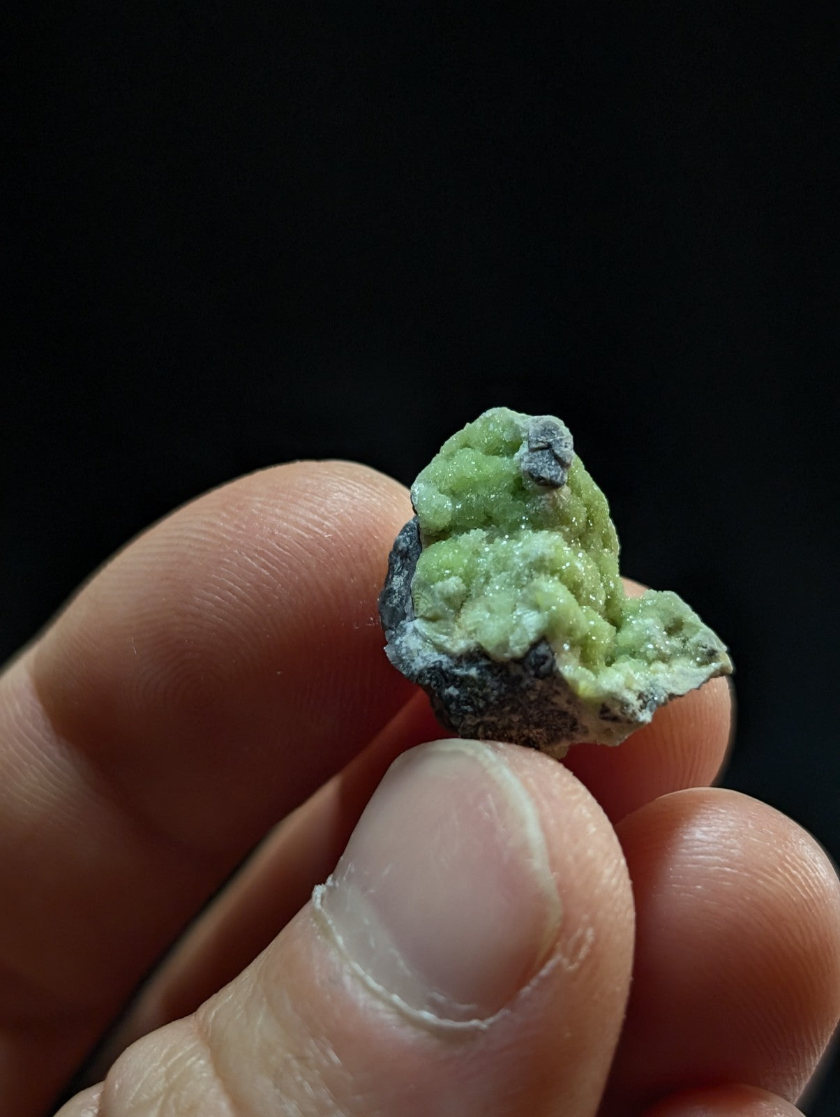 A small, irregularly shaped green mineral specimen with a rough, crystalline texture and dark gray matrix.