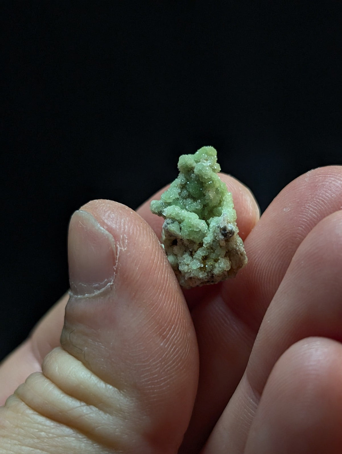 A small, textured green mineral specimen with crystalline formations held between fingers.