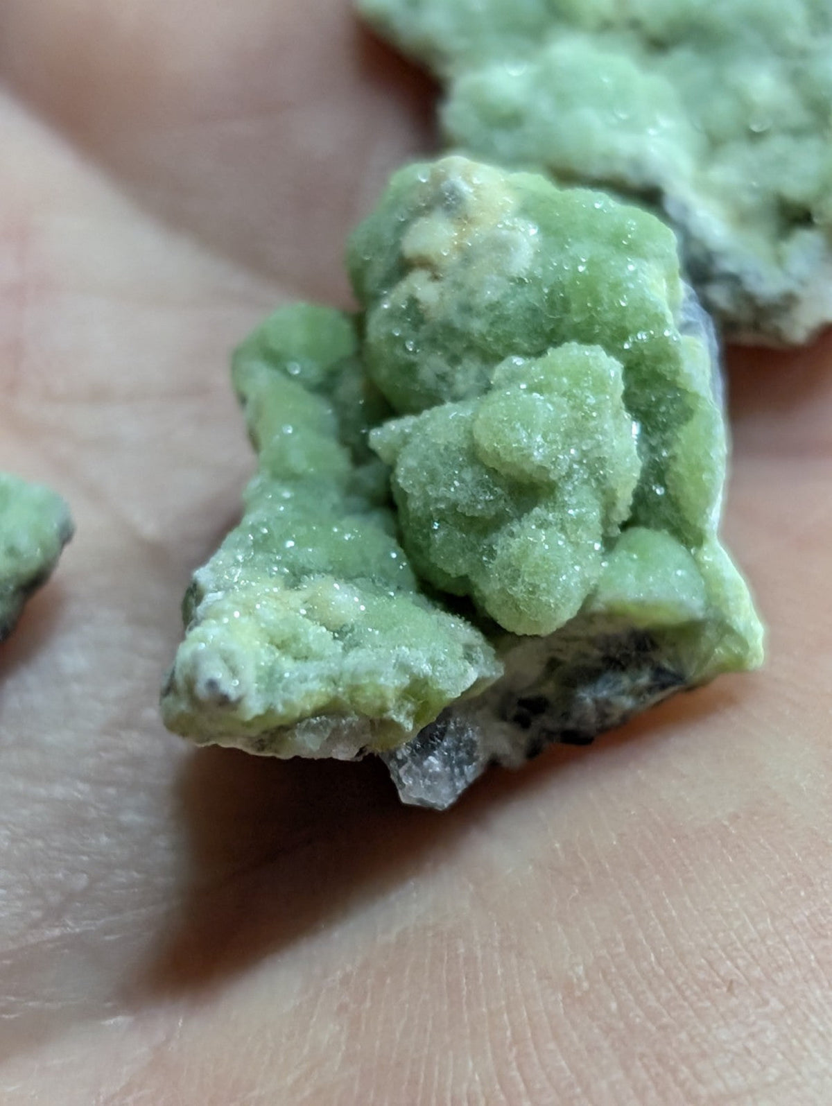 A cluster of light green, crystalline minerals with a rough, textured surface and some darker grayish patches.