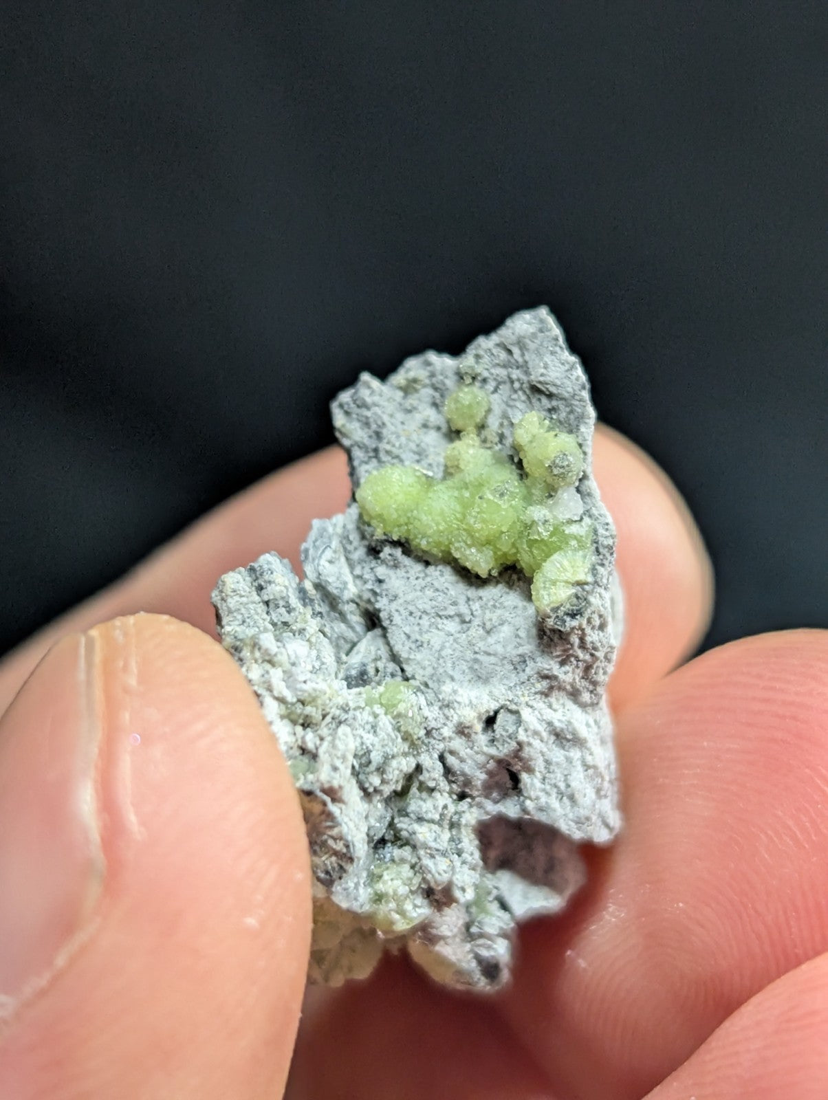 A hand holds a gray, rough-textured mineral specimen featuring small clusters of vibrant green crystals.