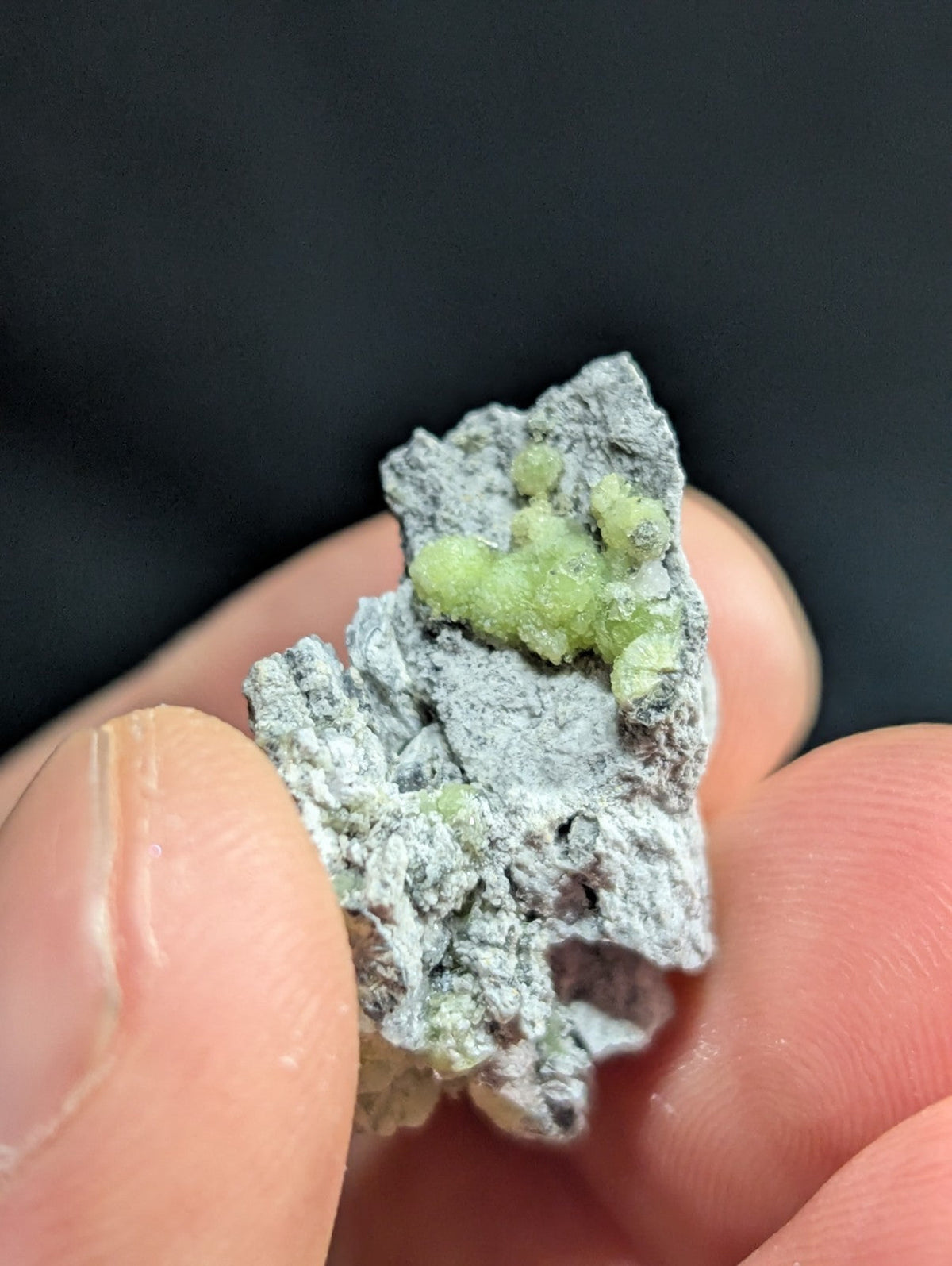 A hand holds a gray, rough-textured mineral specimen featuring small clusters of vibrant green crystals.