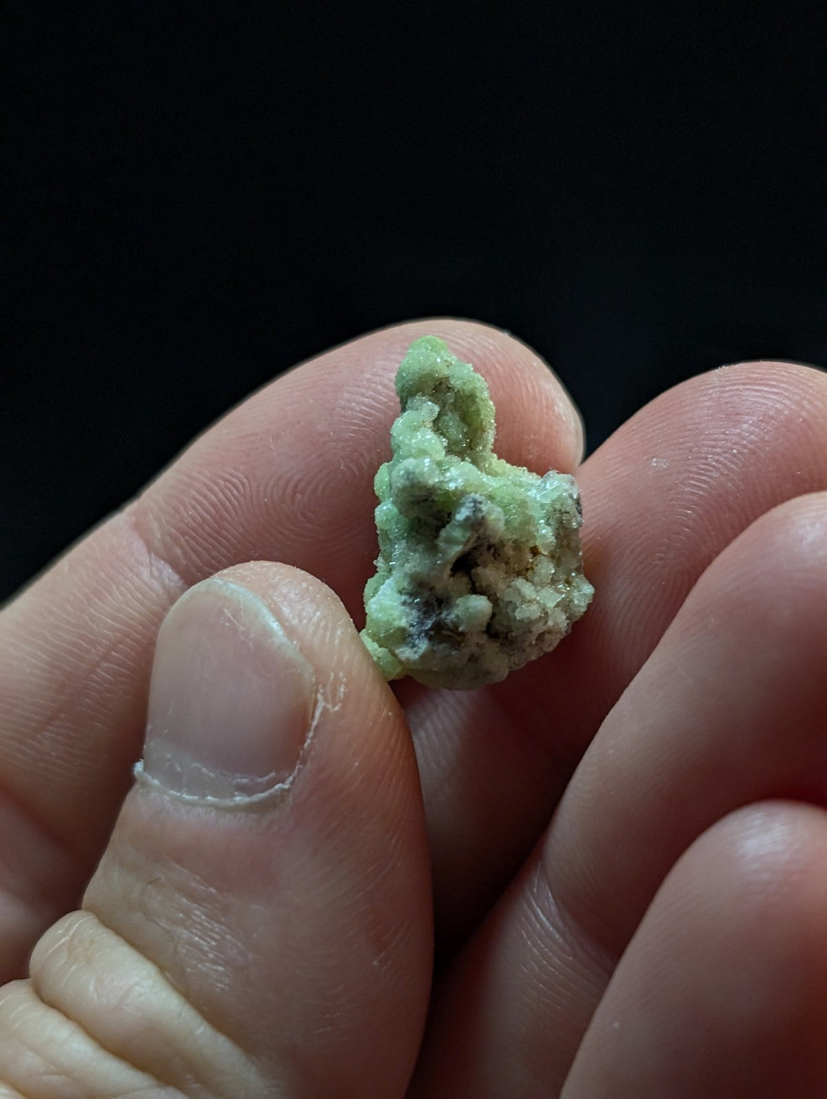 A small, irregularly shaped green mineral specimen with a rough, textured surface and subtle darker patches.