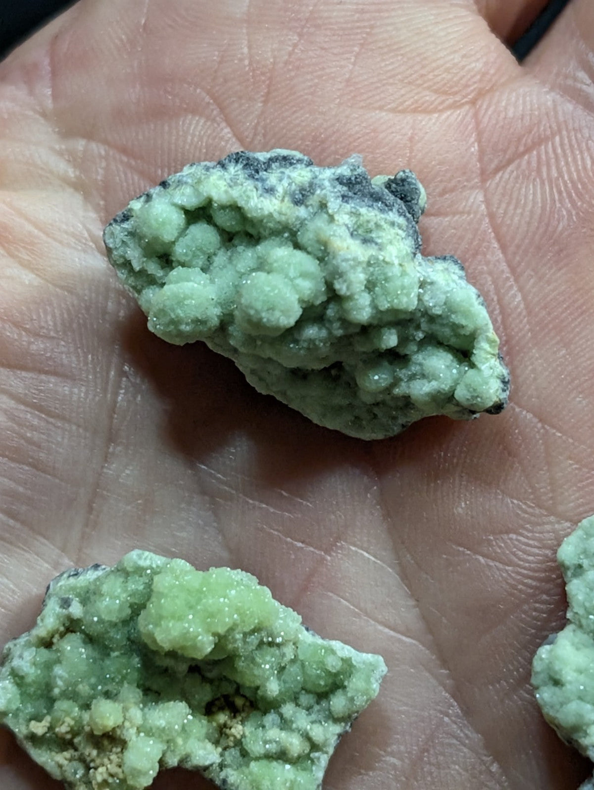 A rough, light green mineral specimen with a crystalline texture and dark gray patches.