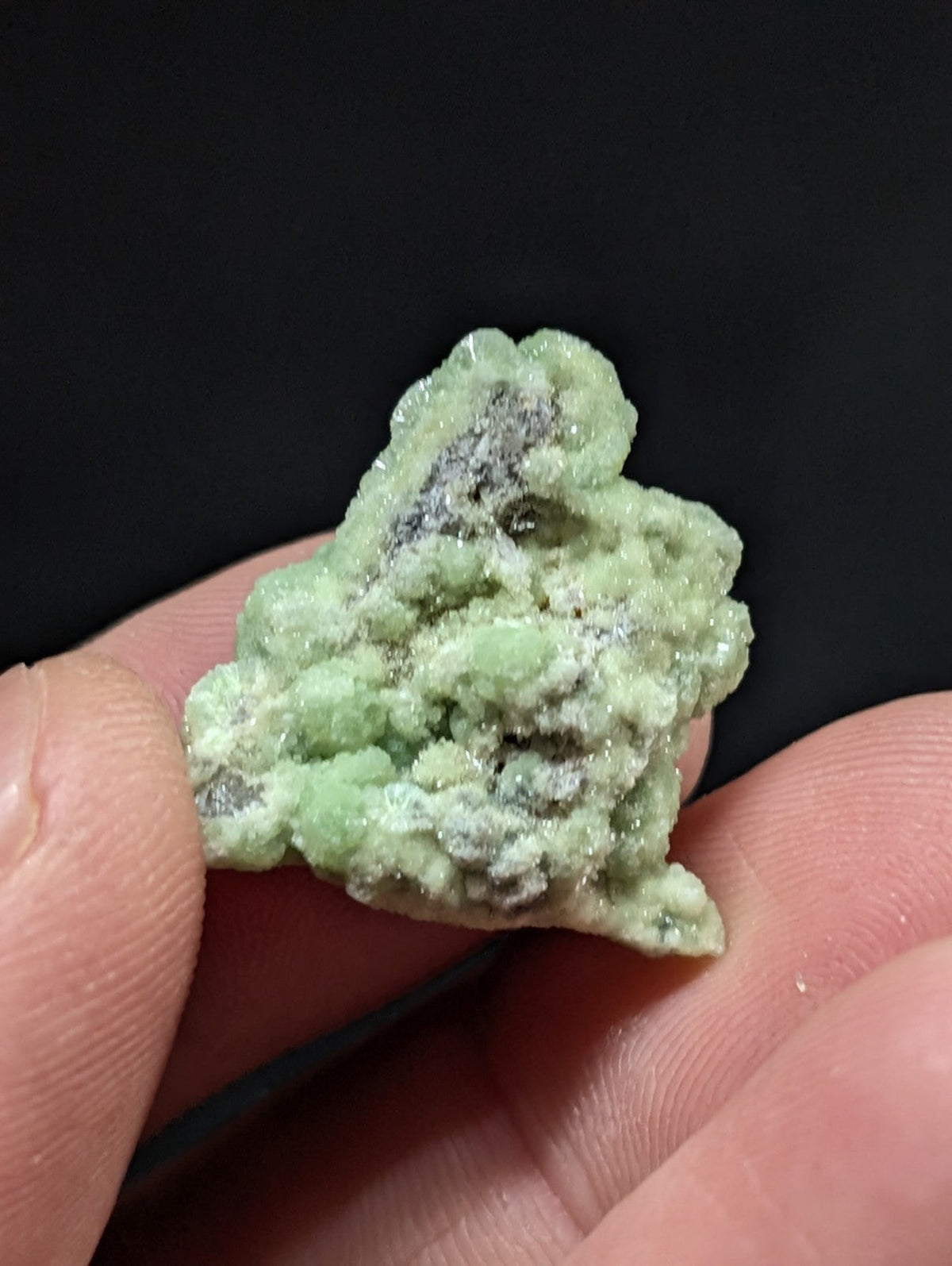 A pale green, crystalline mineral specimen with a rough, textured surface and scattered darker gray inclusions.