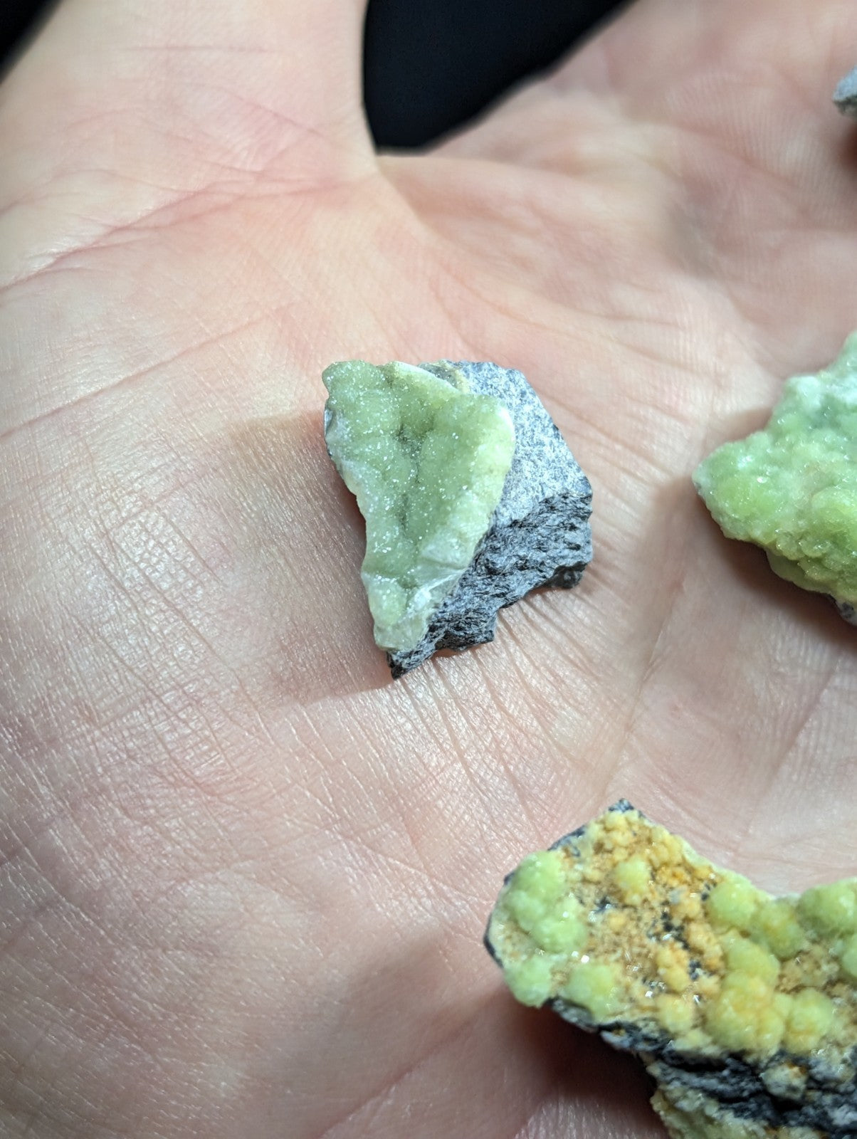 A hand holds a small, irregularly shaped piece of green mineral with a rough texture and crystalline surface.