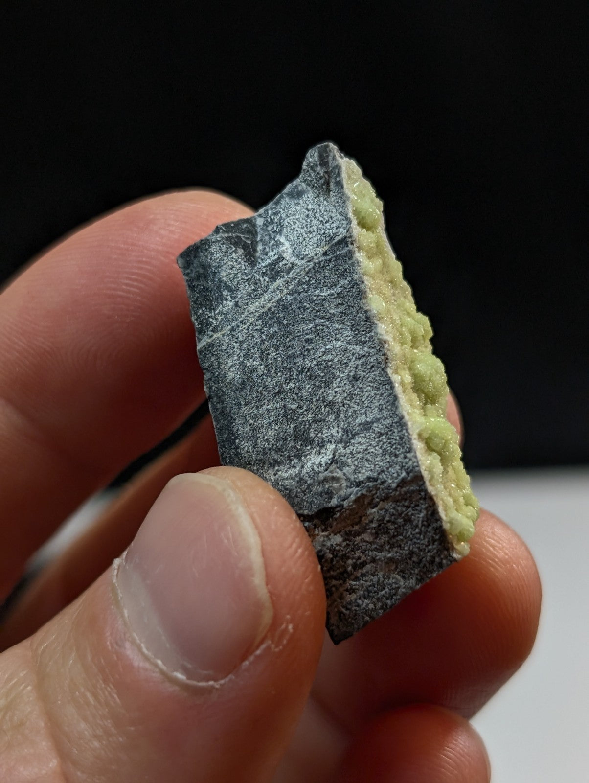A dark gray, rough-textured mineral specimen with a contrasting light green crystalline growth along one edge.