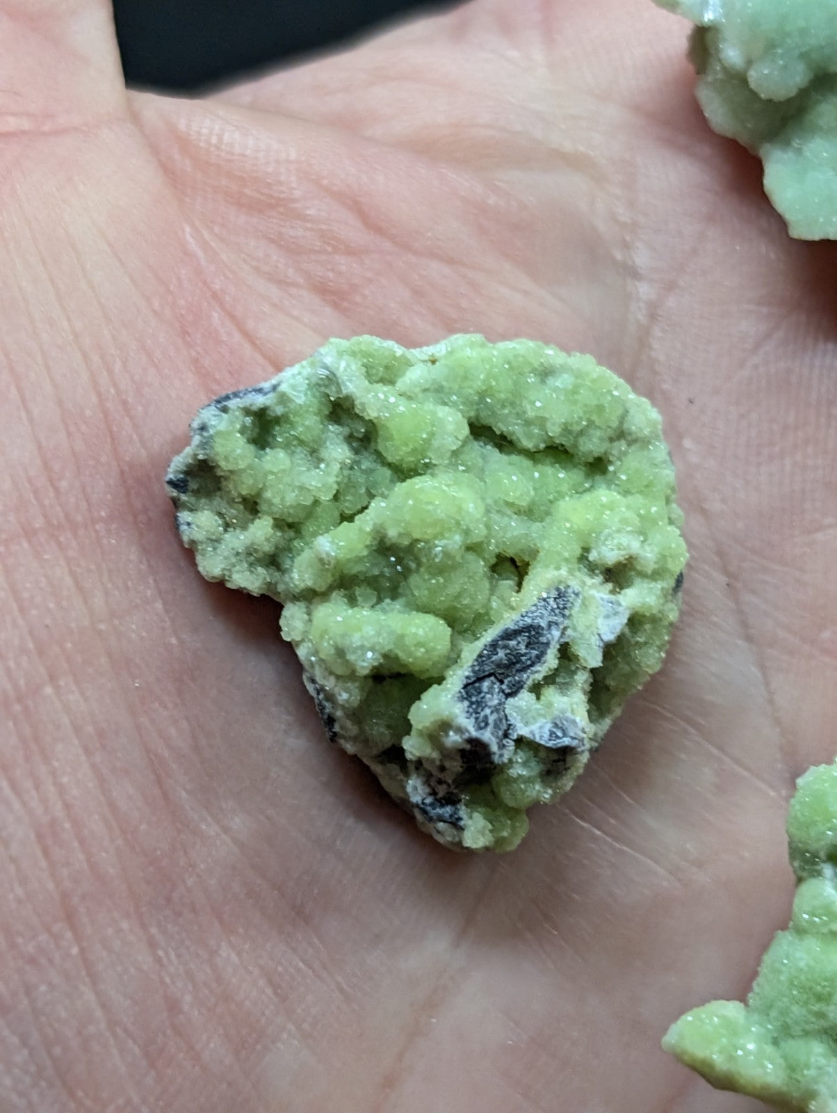 A rough, light green mineral specimen with a crystalline texture and dark gray patches resting in the palm of a hand.