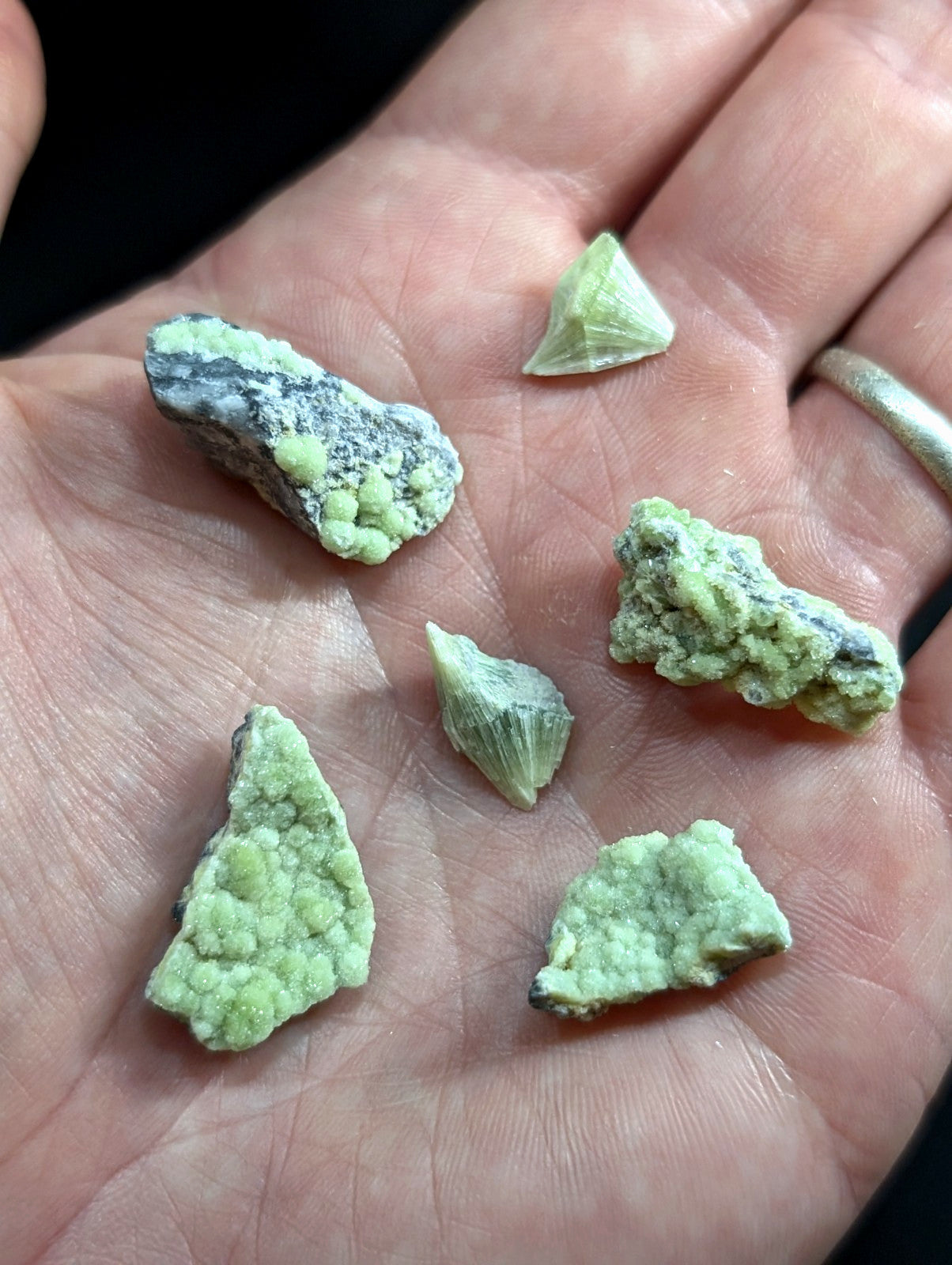 A hand holds several irregularly shaped pieces of greenish-yellow mineral with a rough, textured surface and some darker gray veining.