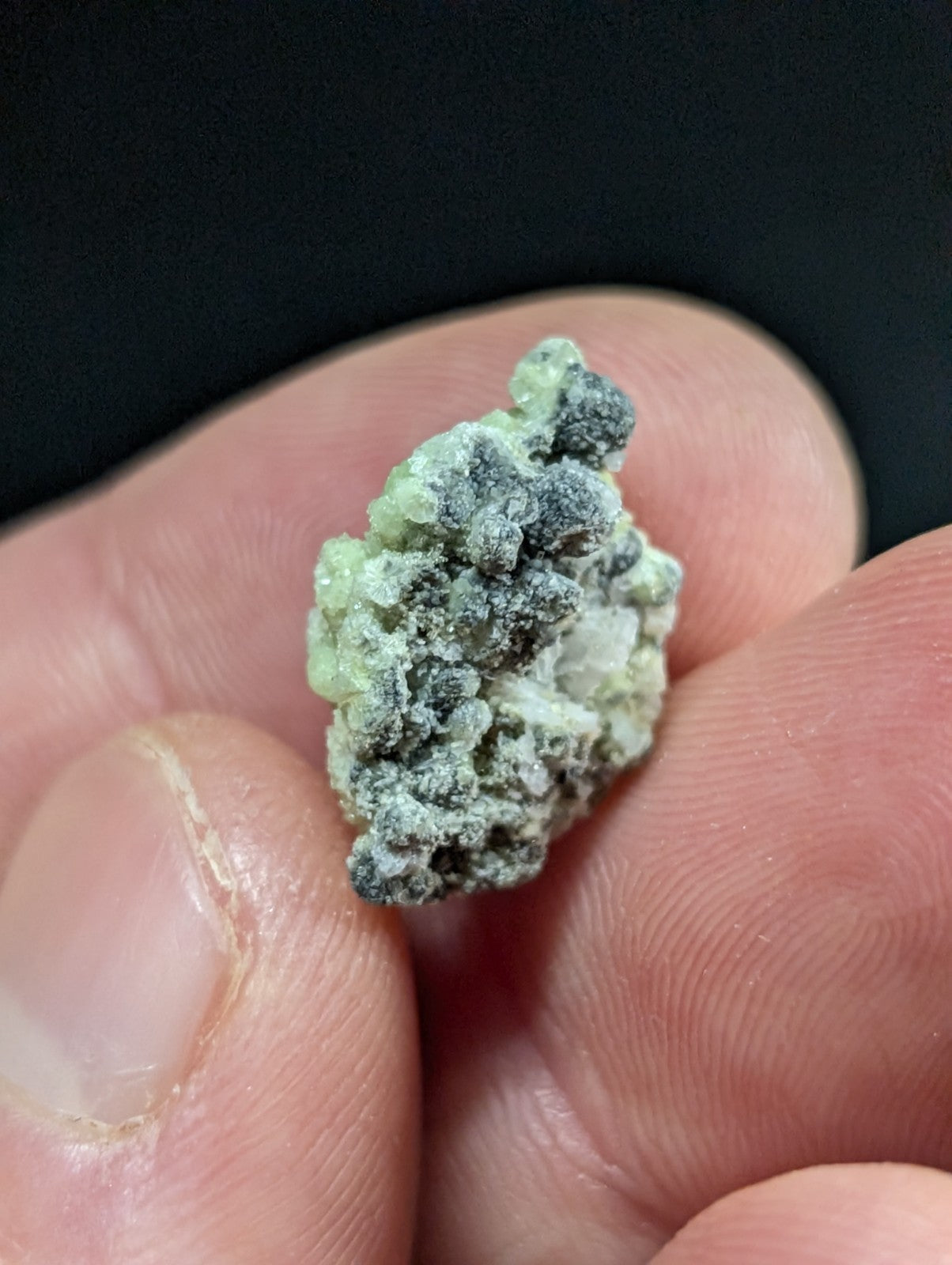 A small, irregularly shaped mineral specimen with a textured surface featuring patches of pale green and dark gray.