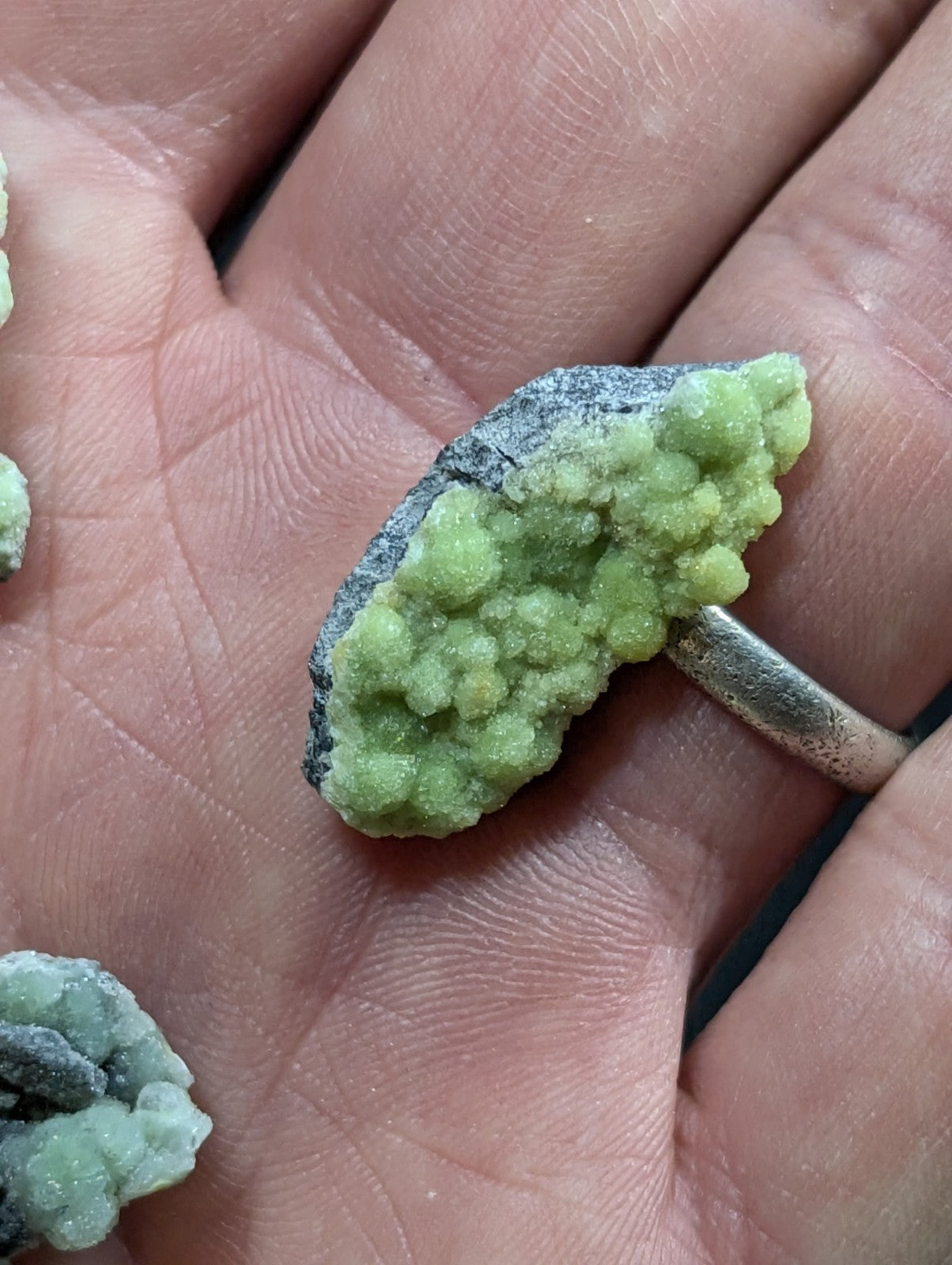 A hand holds a small, irregularly shaped mineral specimen with vibrant green crystalline formations growing from a dark gray base.
