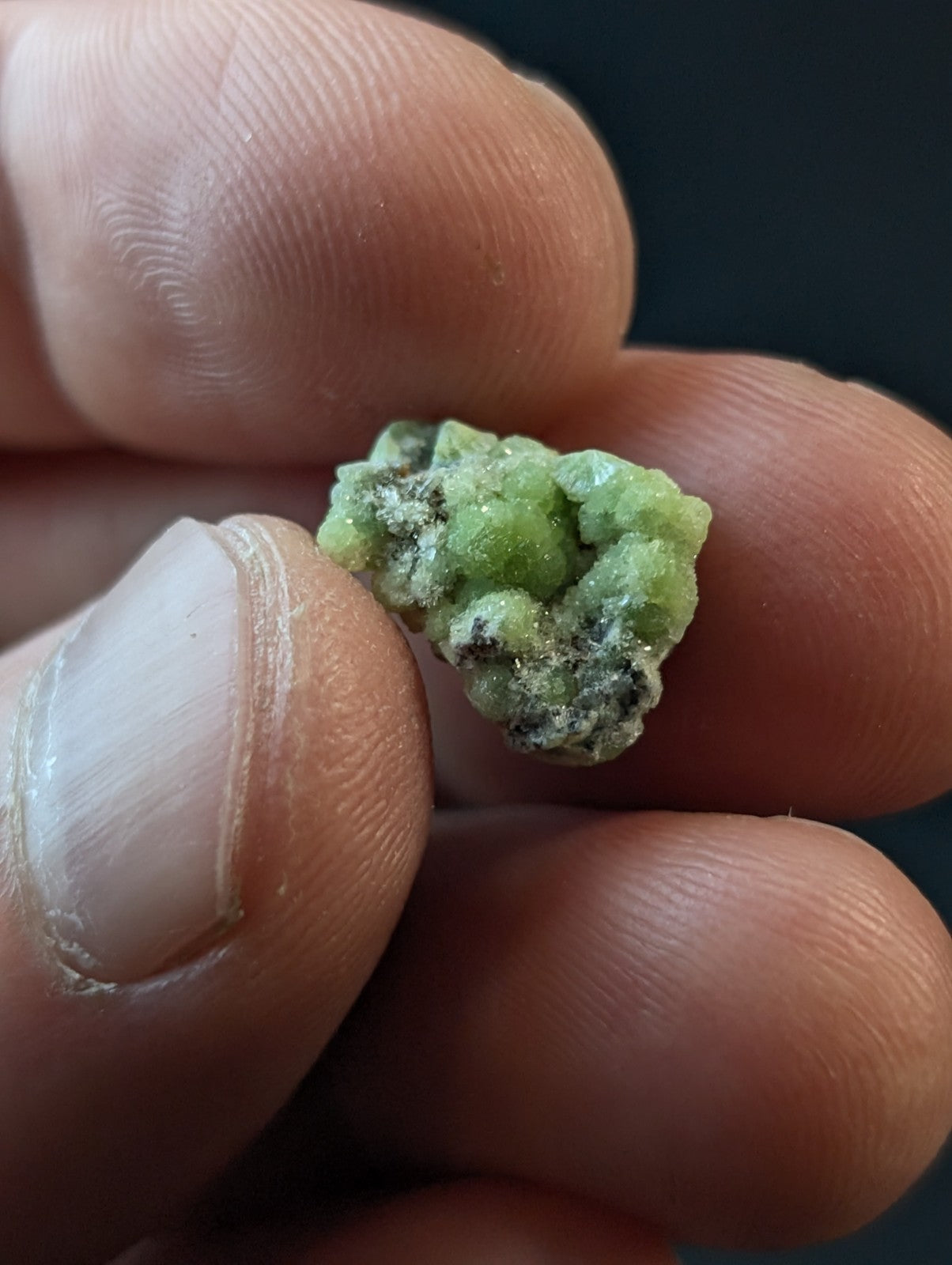 A small, irregularly shaped green mineral specimen with a rough, crystalline texture and patches of dark speckling.