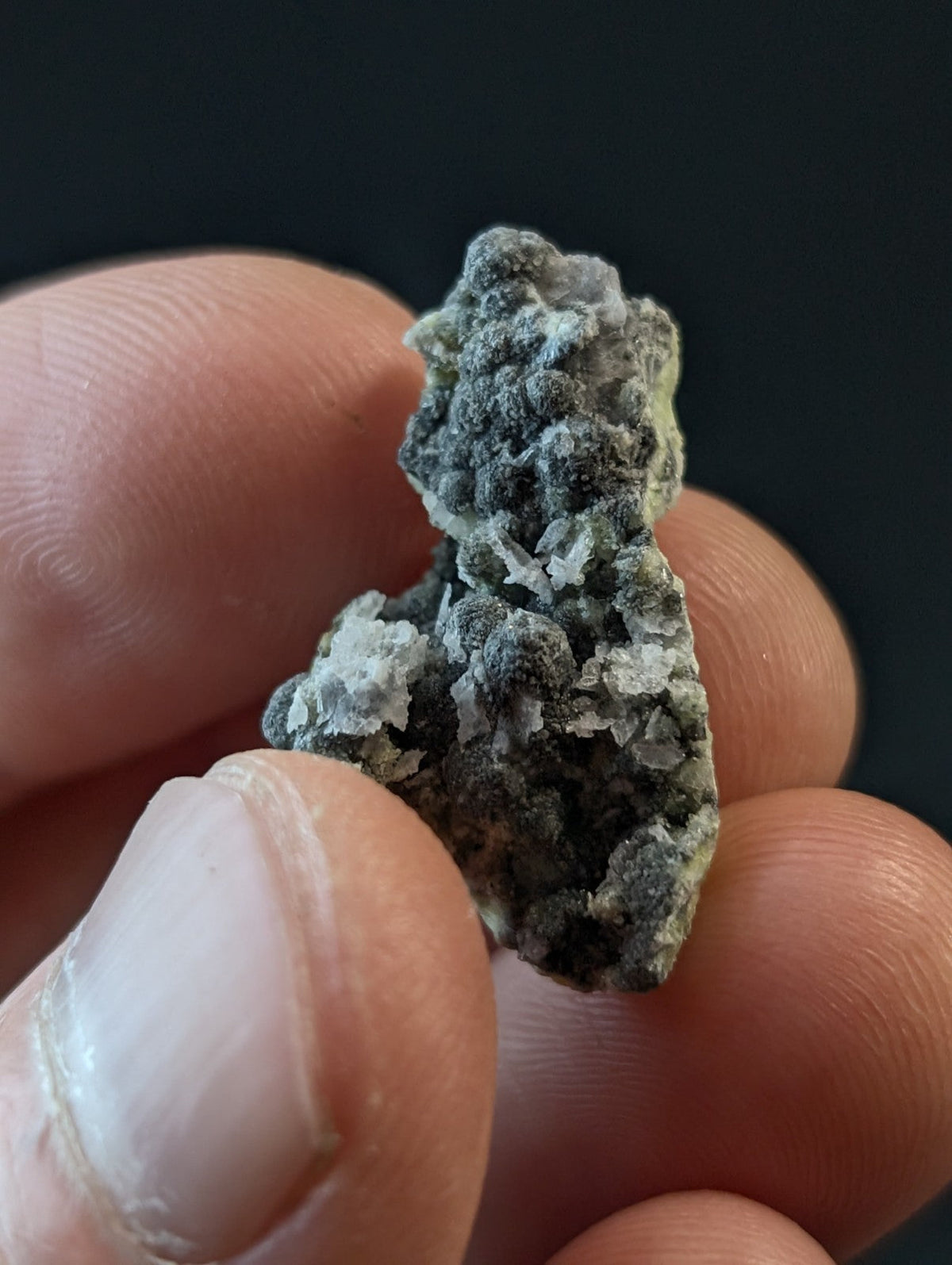 A textured, grayish-green mineral specimen with a rough, crystalline surface held between fingers.