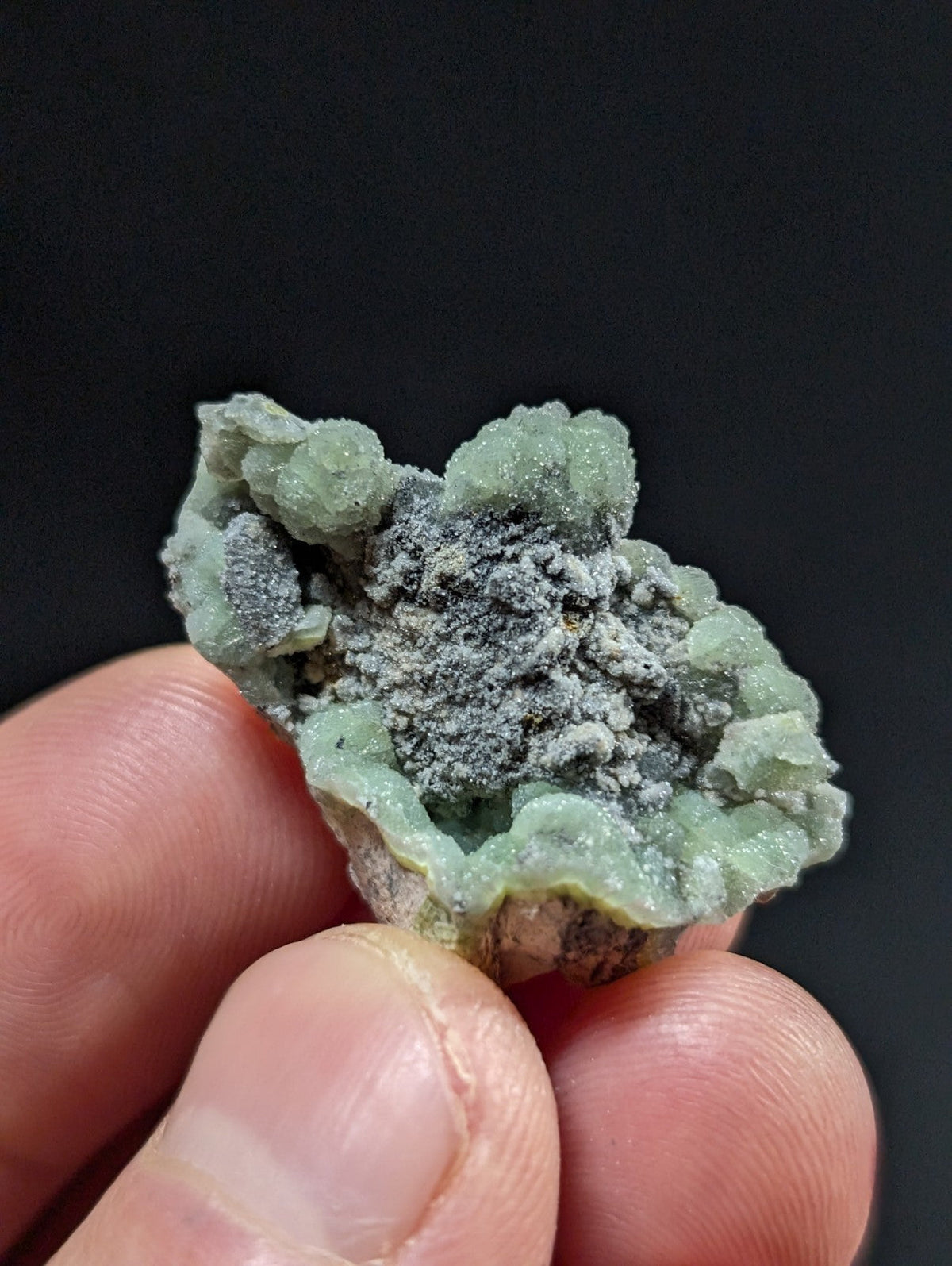 A hand holds a small, irregularly shaped mineral specimen featuring translucent green crystalline formations with a central cluster of grayish powder.