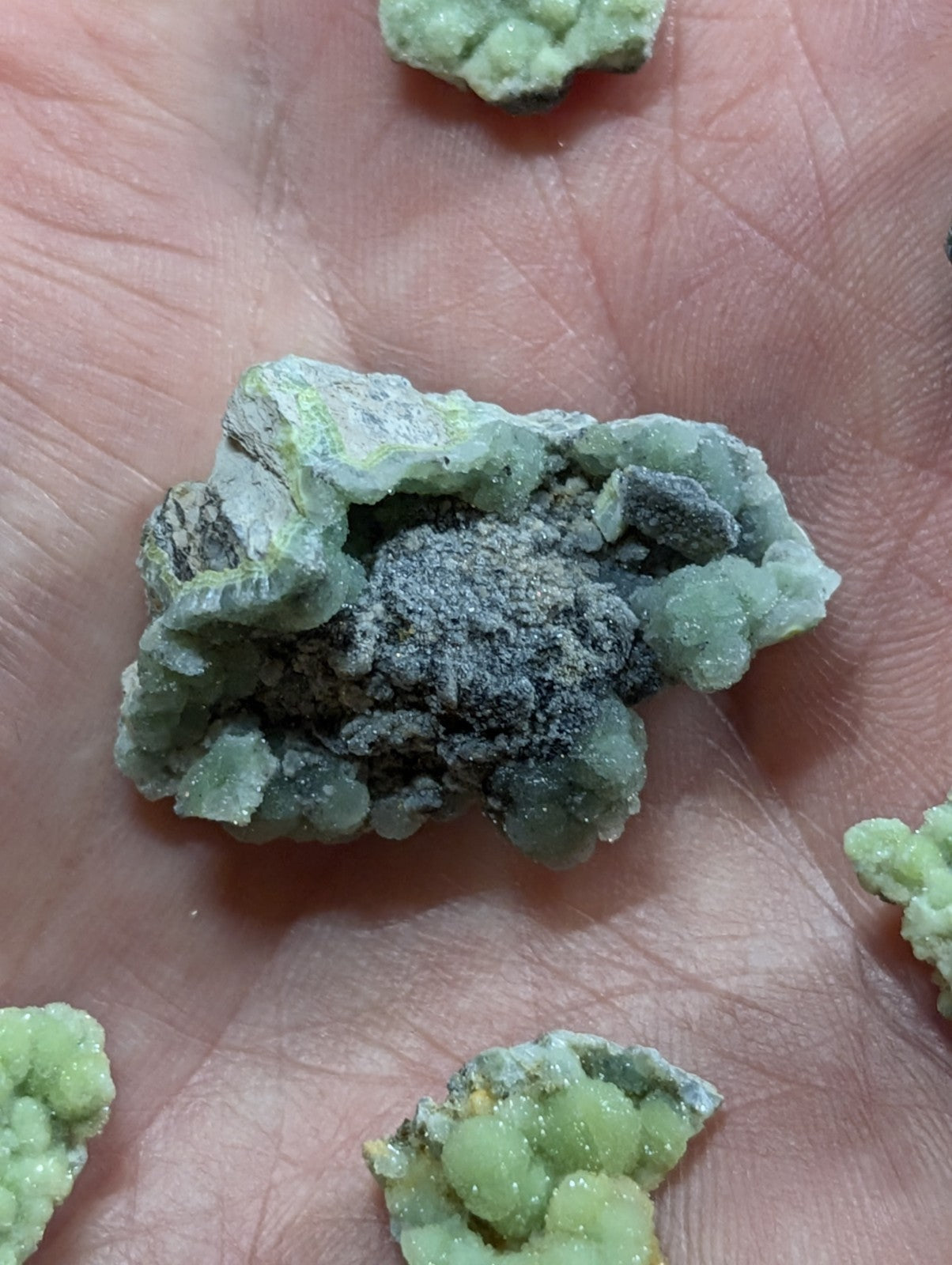 A rough, pale green mineral specimen with a crystalline texture and dark grayish-black inclusions.