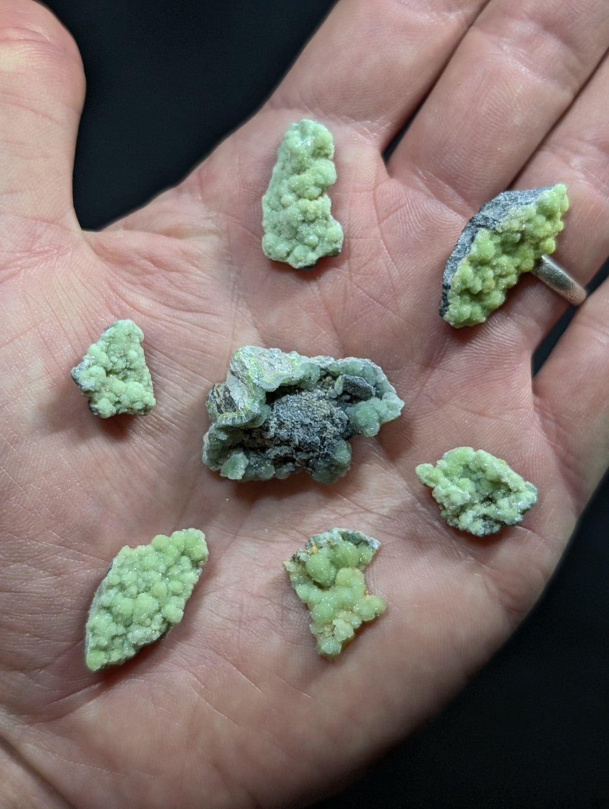 A hand holds several irregularly shaped, light green mineral specimens with a textured, crystalline surface and some darker grayish patches.
