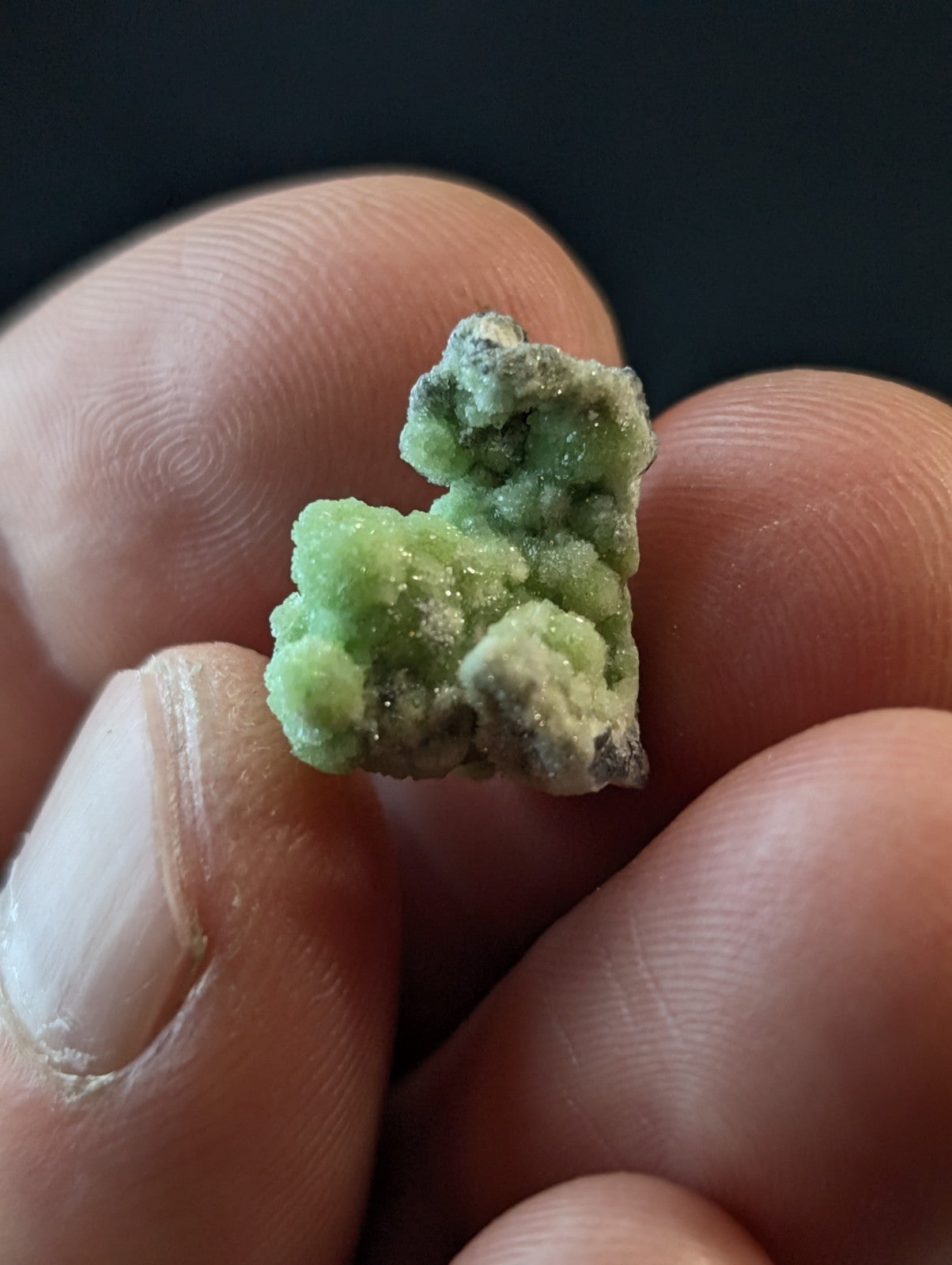 A small, irregularly shaped green mineral specimen with a crystalline texture and some darker grayish patches.