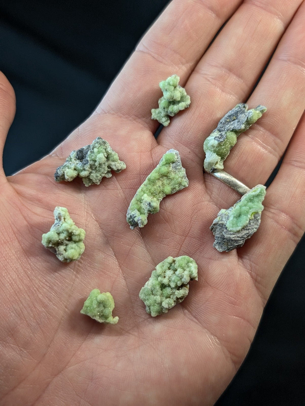 A hand holds several irregularly shaped, light green and grayish mineral specimens with a rough, crystalline texture.