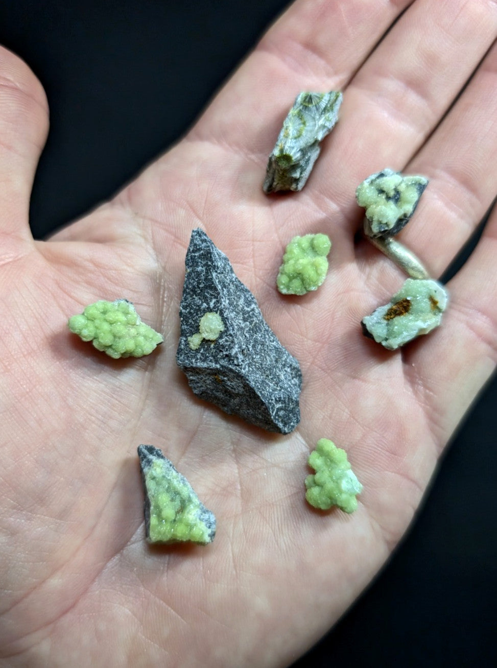 A hand holds a collection of small, irregularly shaped green and gray mineral specimens with crystalline textures.