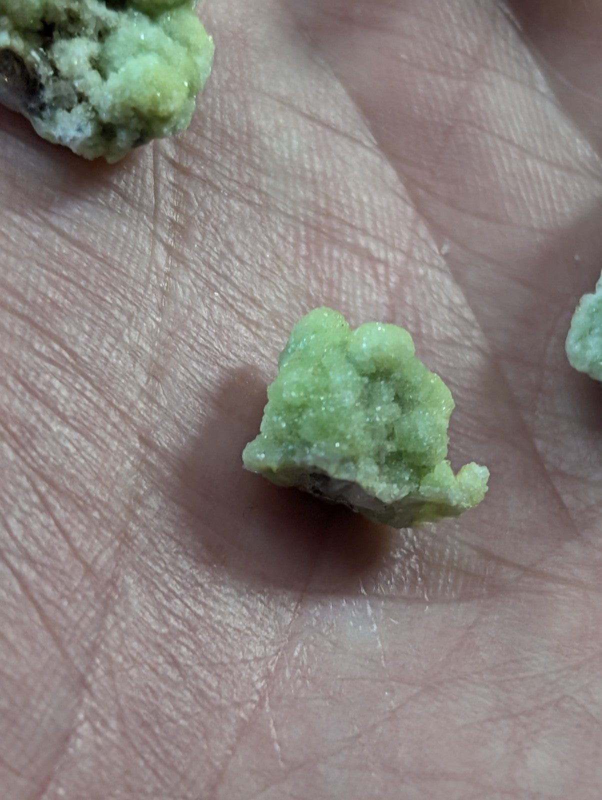 A small, irregularly shaped green mineral specimen with a rough, crystalline texture resting in the palm of a hand.