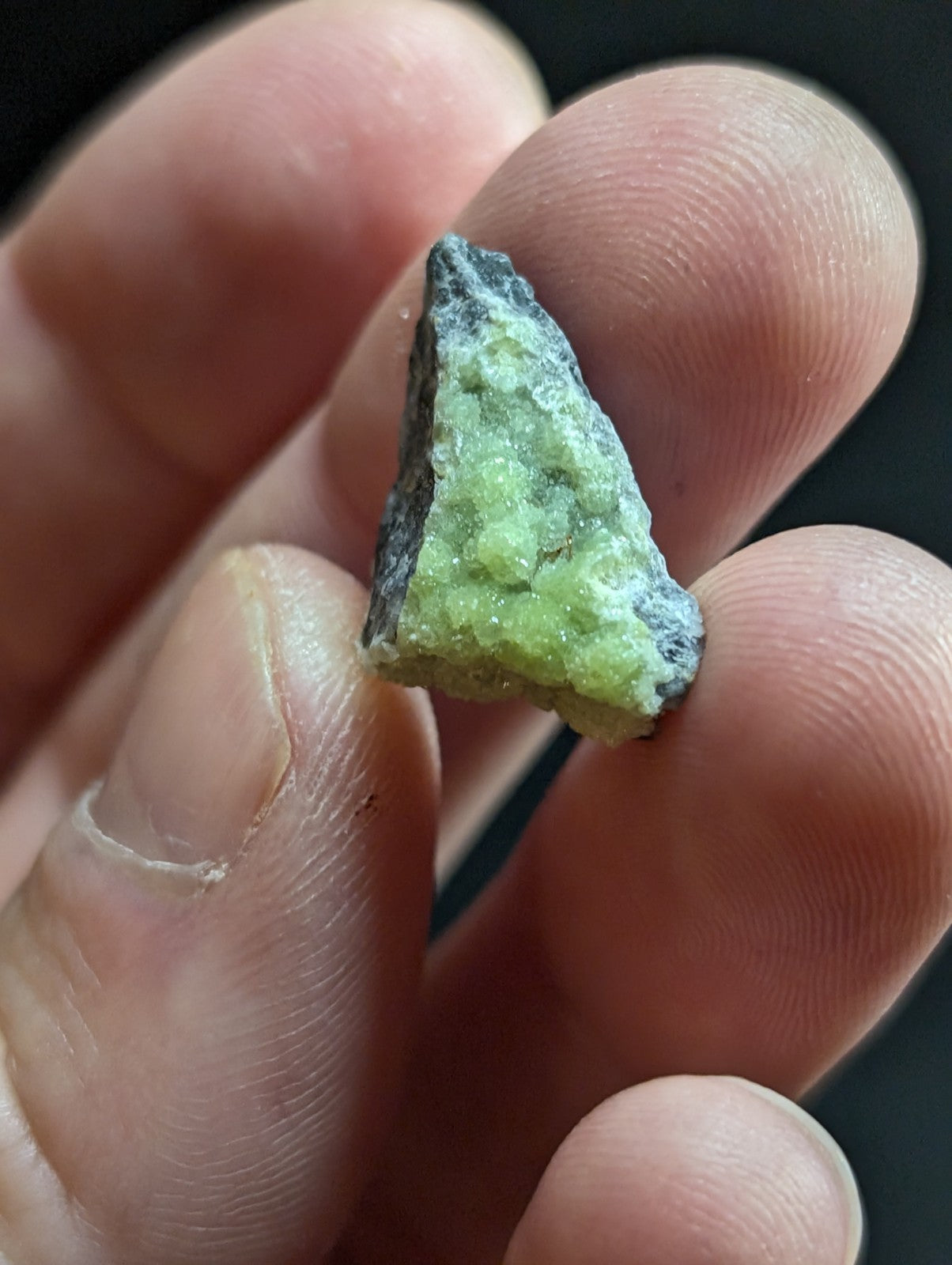 A triangular, rough-textured green mineral specimen with a darker grayish-black crust along its edges.