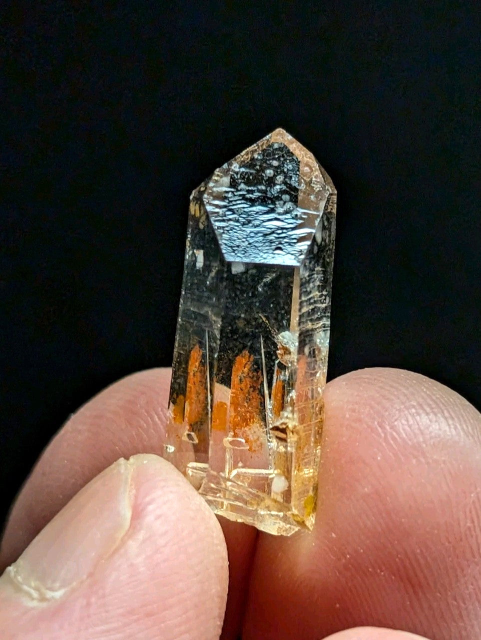 A translucent yellowish quartz crystal with a pointed top and internal orange inclusions, held between fingers against a dark background.