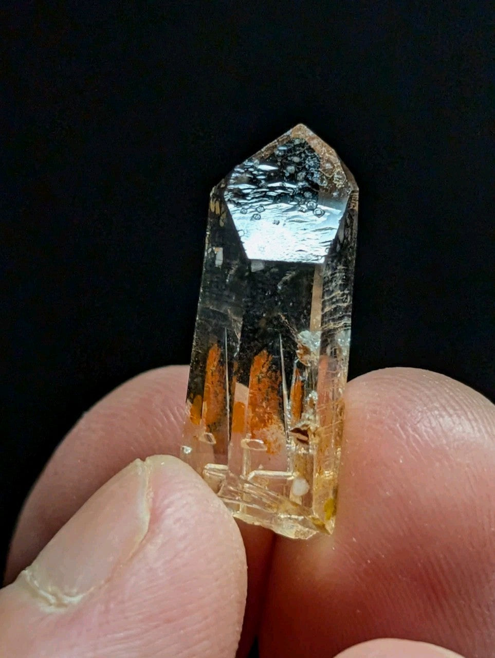 A translucent yellowish quartz crystal with a pointed top and visible internal orange inclusions, held between fingers.