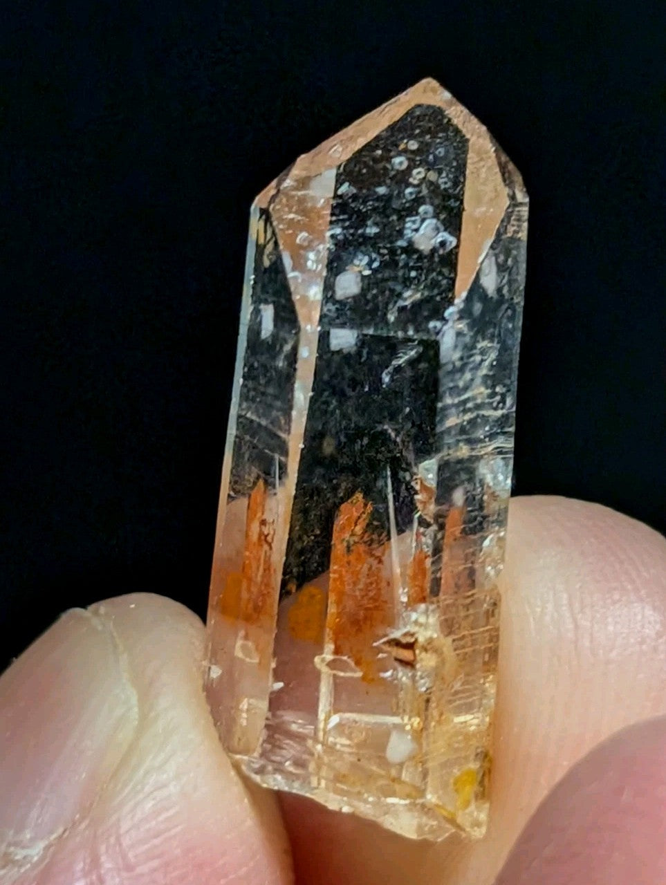 A translucent orange and clear quartz crystal with dark inclusions, held between fingers.