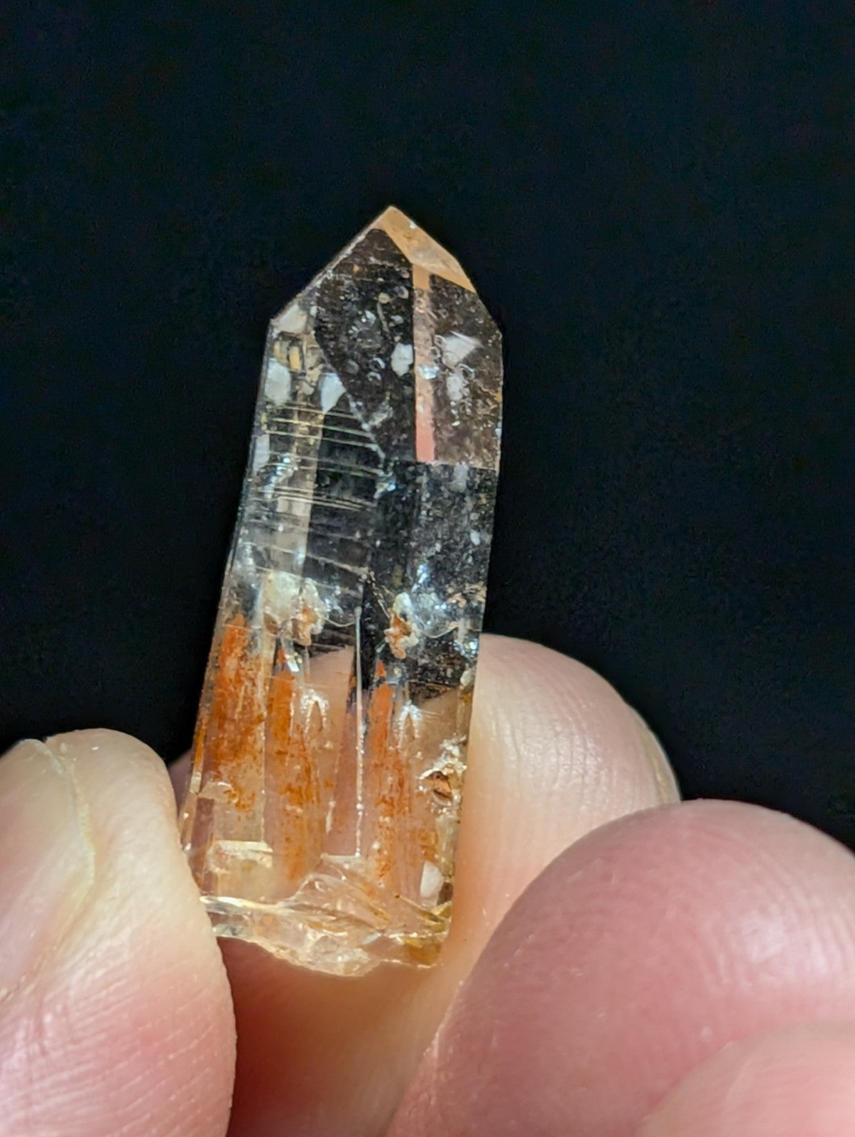 A translucent orange and clear quartz crystal with natural inclusions, held between fingers against a dark background.