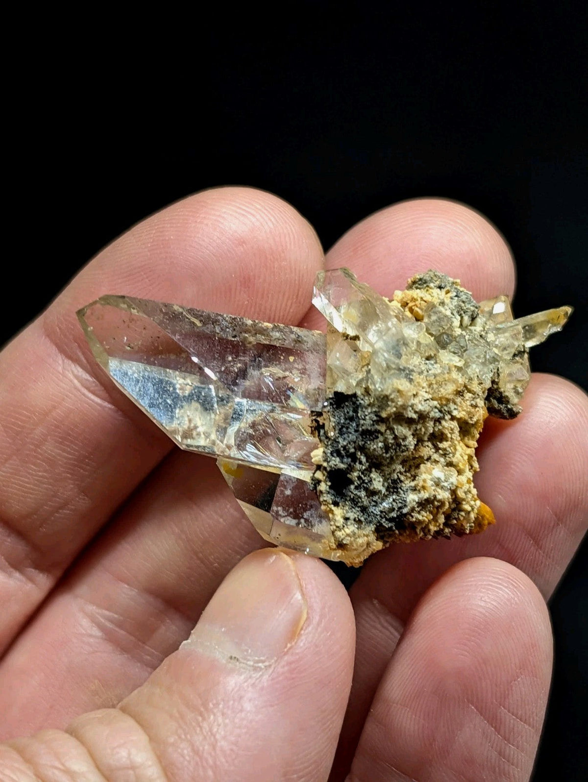 A translucent, clear quartz crystal with sharp facets and a natural matrix of earthy yellow and dark green minerals.