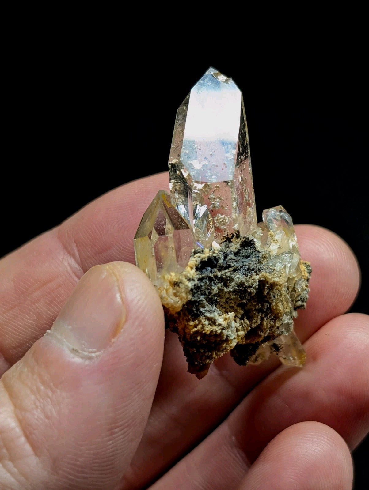A clear quartz crystal cluster with a prominent central point, attached to a rough, earthy matrix of dark green and yellowish mineral material.