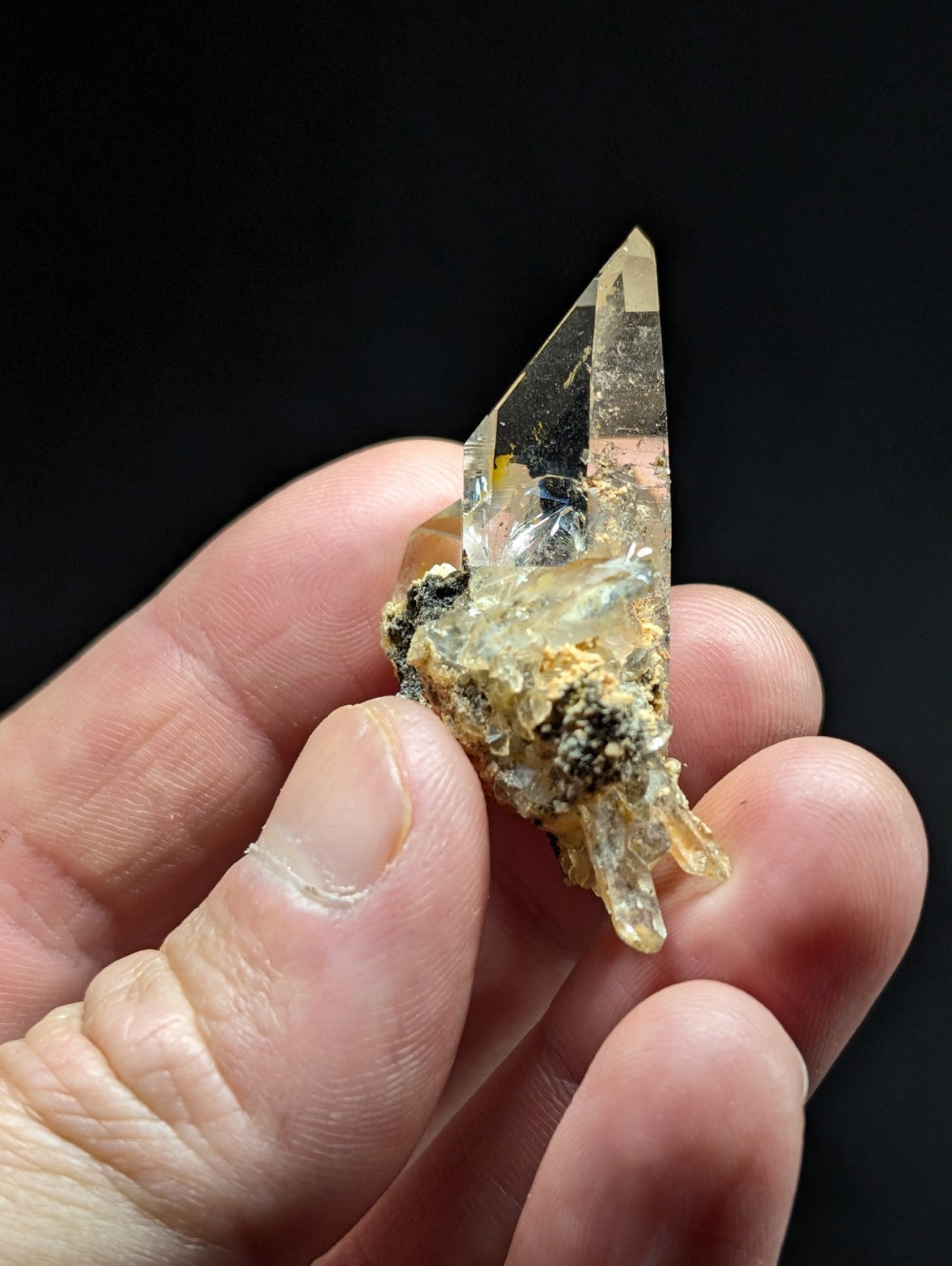 A translucent, pointed quartz crystal with a clear and slightly yellowish hue, embedded in a rough, earthy matrix of dark and light mineral deposits.