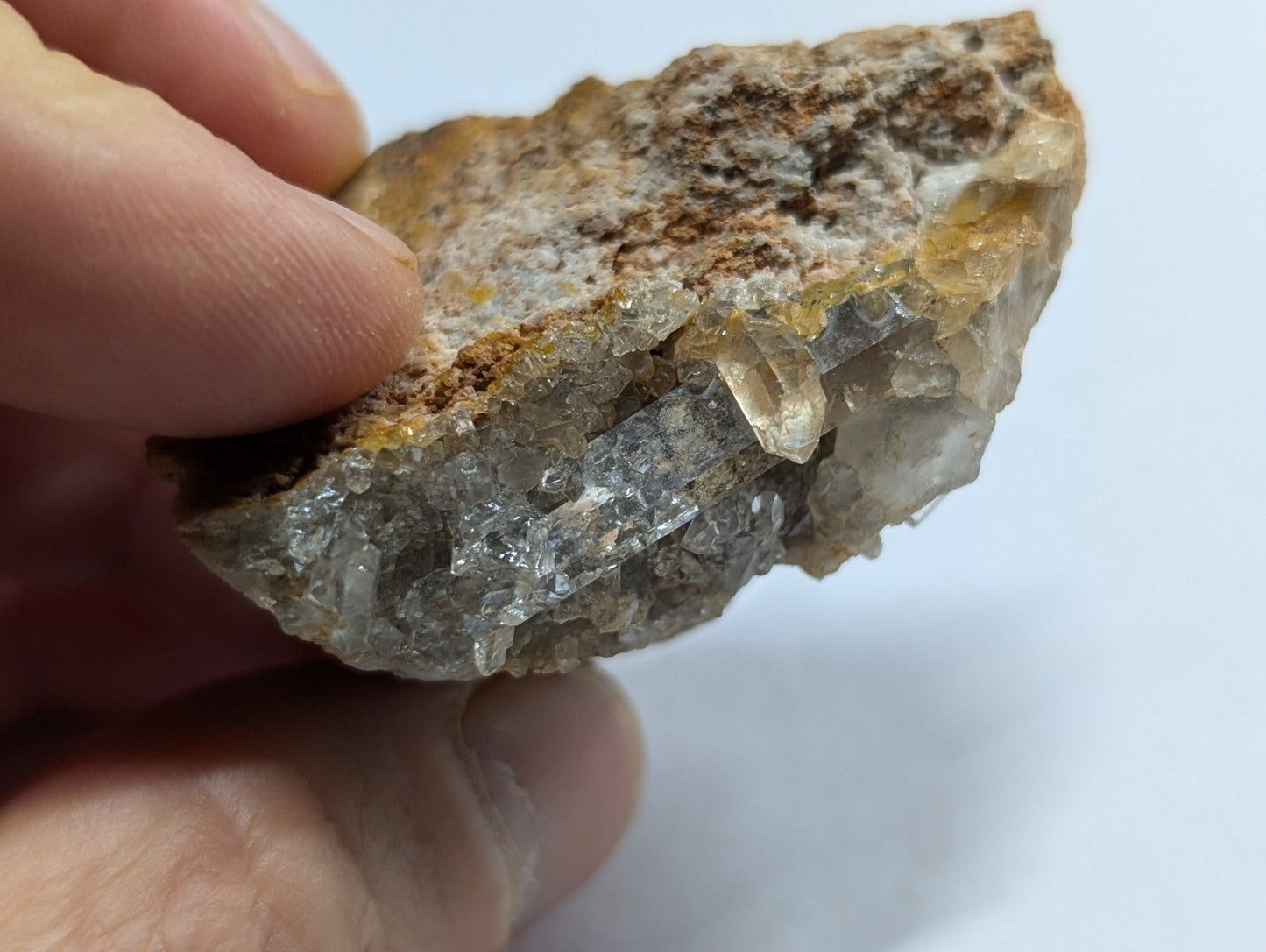 A hand holds a rough, irregularly shaped rock specimen featuring translucent quartz crystals embedded in a matrix of brownish-yellow mineral.
