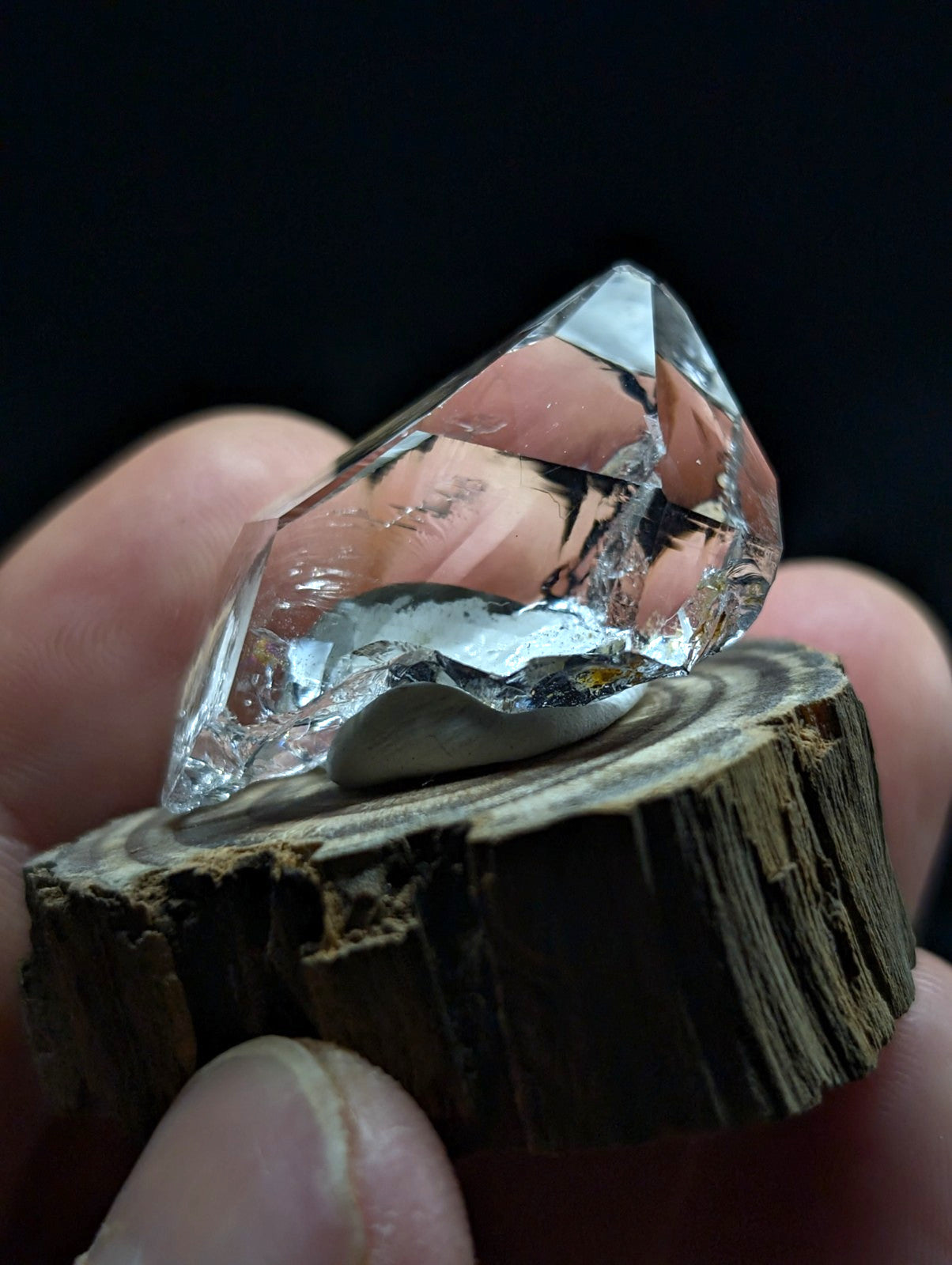A clear, faceted quartz crystal with a smooth, polished base rests on a rustic wooden slice.