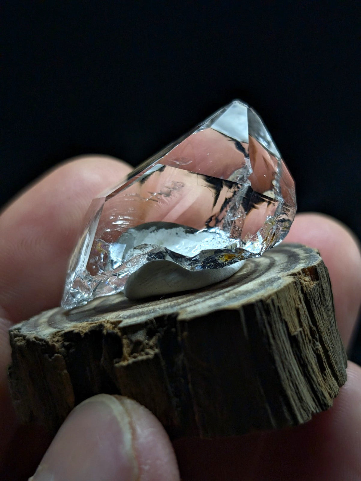A clear, faceted quartz crystal with a smooth, polished base rests on a rustic wooden slice.