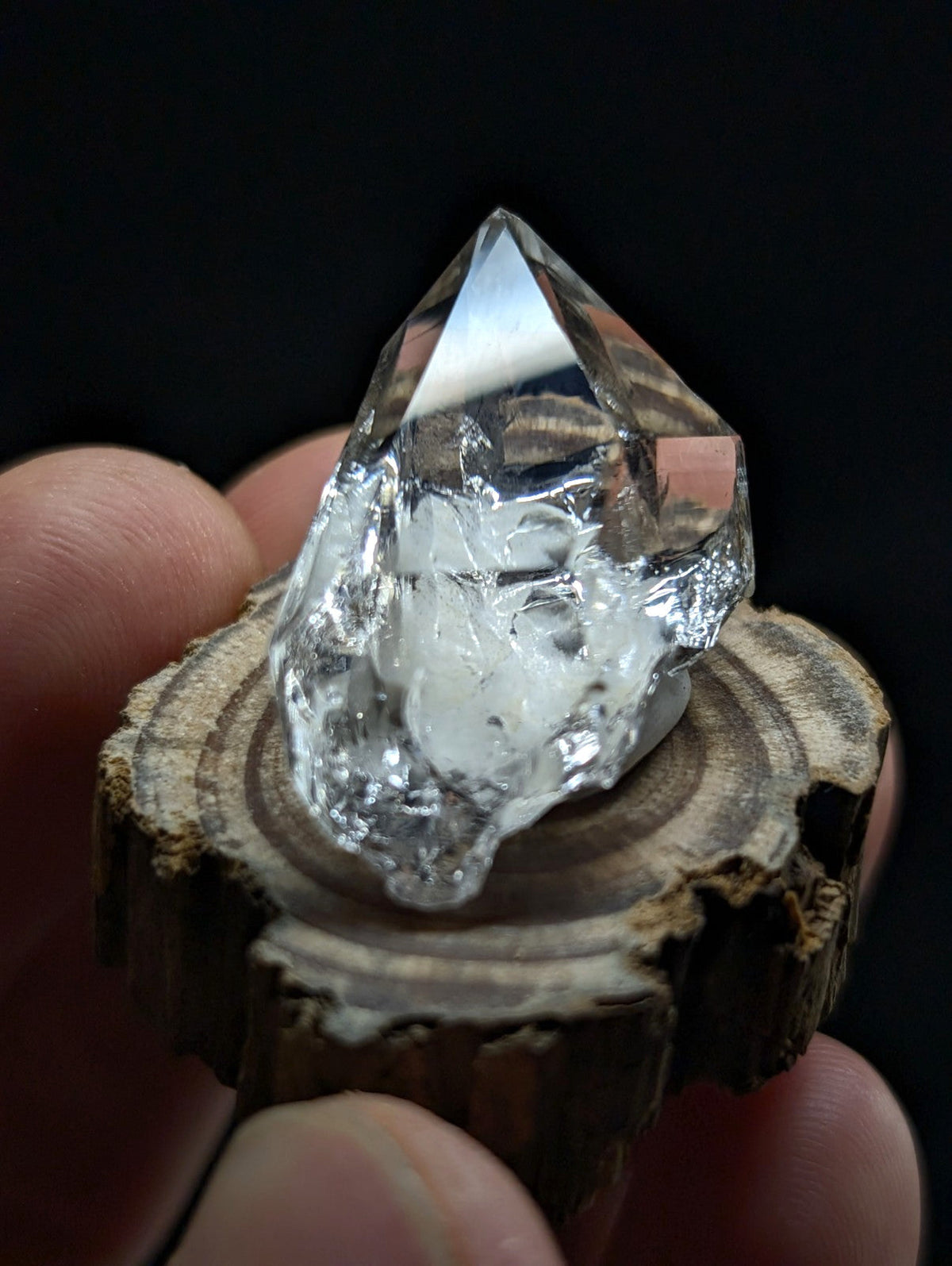 A clear, faceted quartz crystal with natural inclusions and a pointed tip rests on a rustic wooden slice.