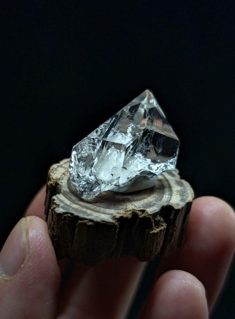 A clear, faceted quartz crystal with a pointed tip and natural internal inclusions rests on a rustic wooden slice.