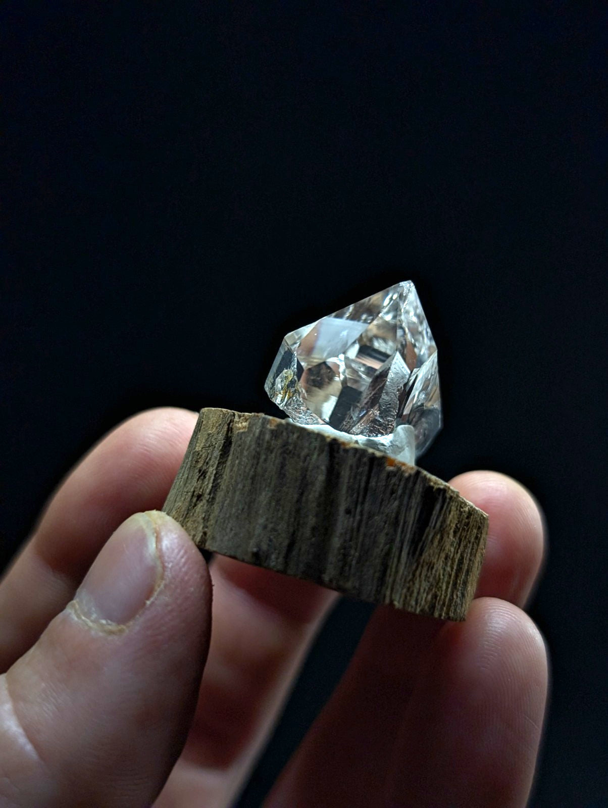 A clear, faceted crystal gemstone rests atop a rustic wooden slice held in a hand.