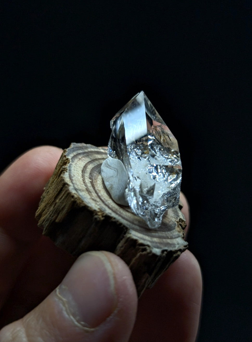 A clear, faceted quartz crystal with a metallic sheen sits atop a rustic wooden slice held in someone’s hand.