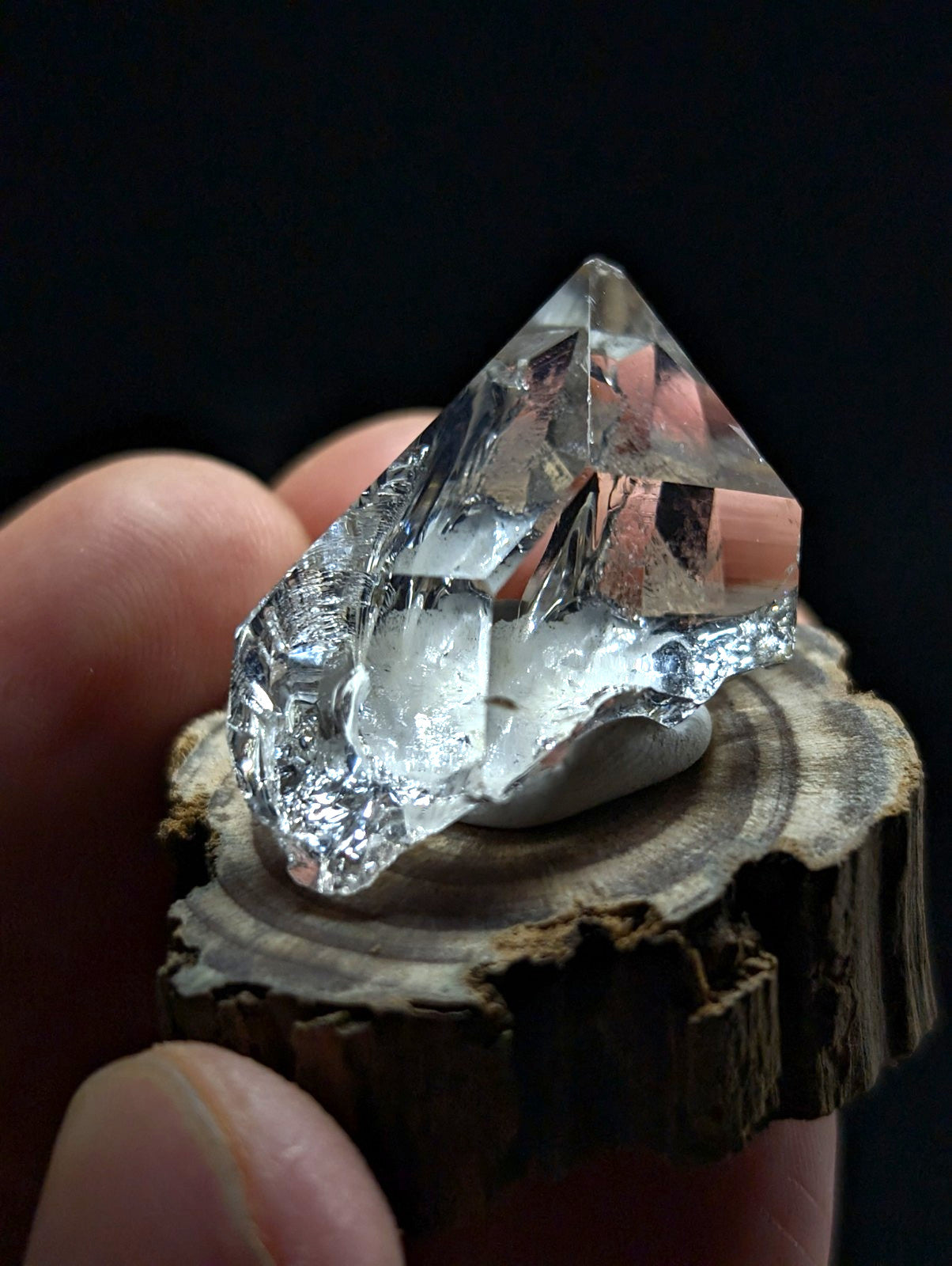 A clear, faceted quartz crystal with a pointed tip and natural imperfections rests on a rustic wooden slice.