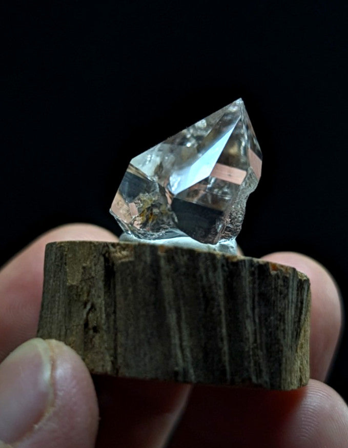 A clear, faceted quartz crystal with natural inclusions sits atop a rustic wooden base.