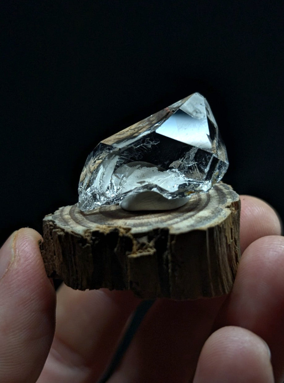 A clear, faceted quartz crystal with a natural, rough-edged base rests on a rustic wooden slice.