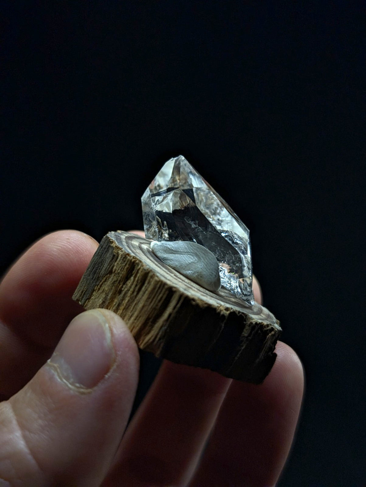 A clear, faceted quartz crystal with a smooth gray stone base rests on a rustic wooden slice.
