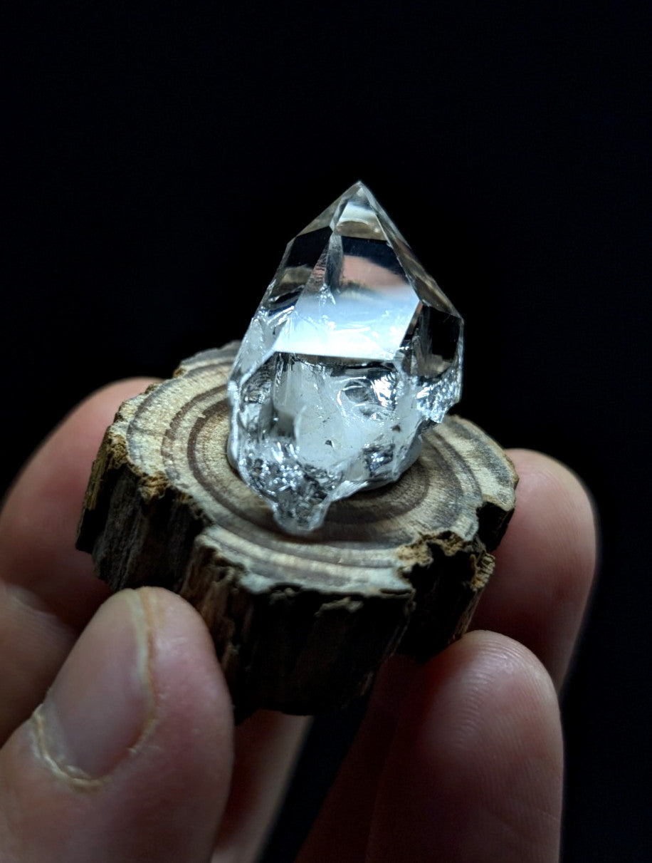 A clear, faceted quartz crystal with a pointed tip and natural inclusions sits atop a rustic wooden slice.