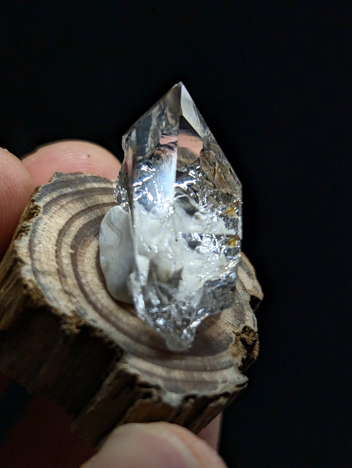 A clear, faceted quartz crystal with a pointed tip and internal inclusions rests on a rustic wooden slice.