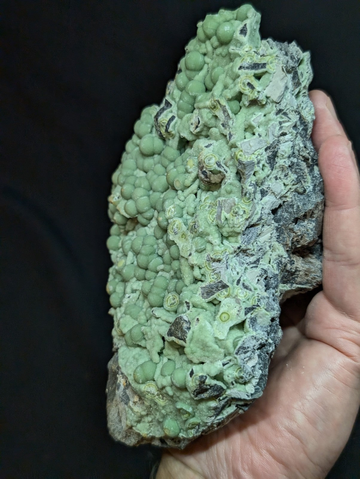 A hand holds a green, bumpy mineral specimen with rounded nodules and patches of darker rock.