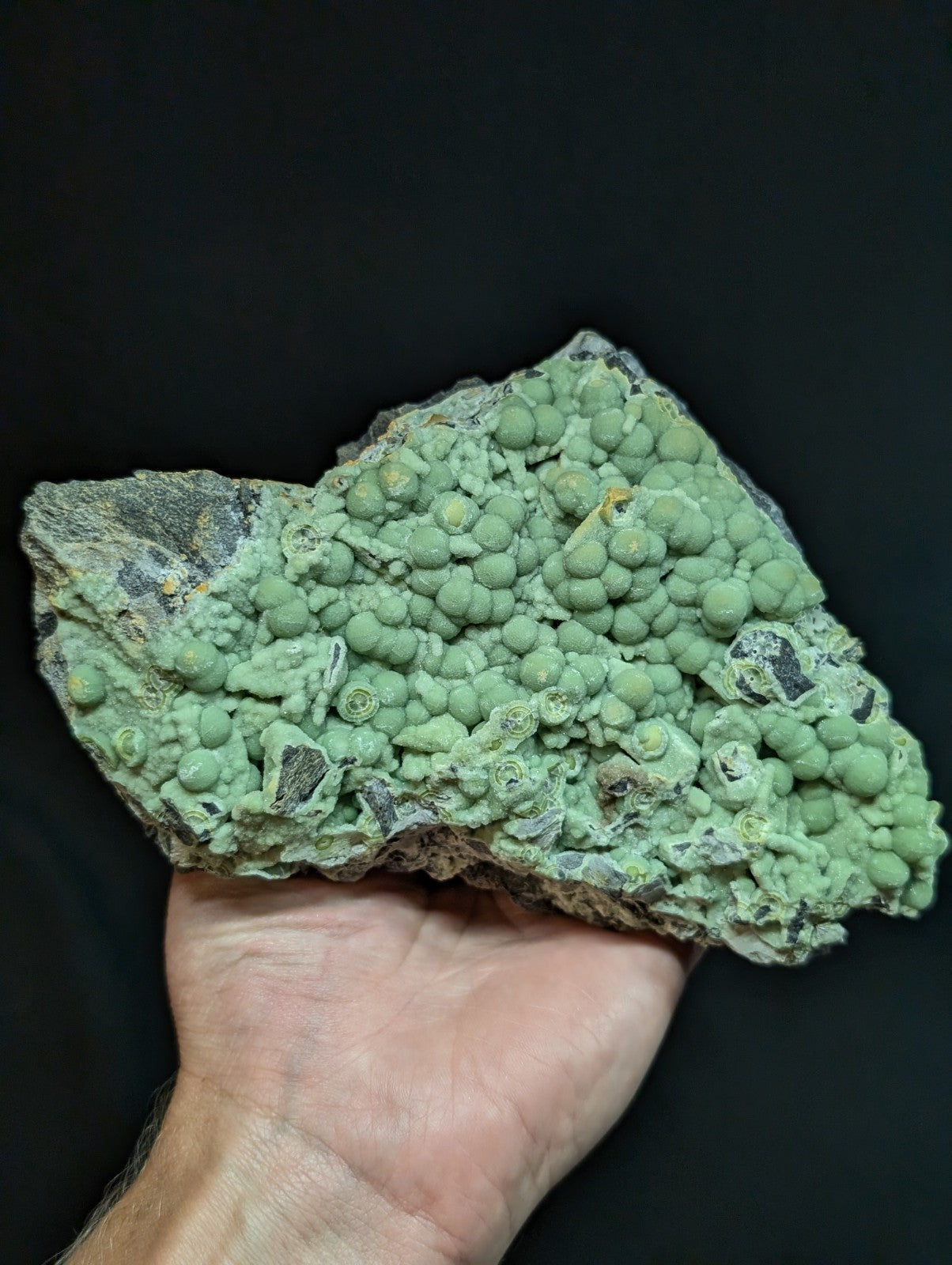 A hand holds a rough, irregularly shaped mineral specimen featuring clusters of small, rounded green nodules with some darker grayish patches.