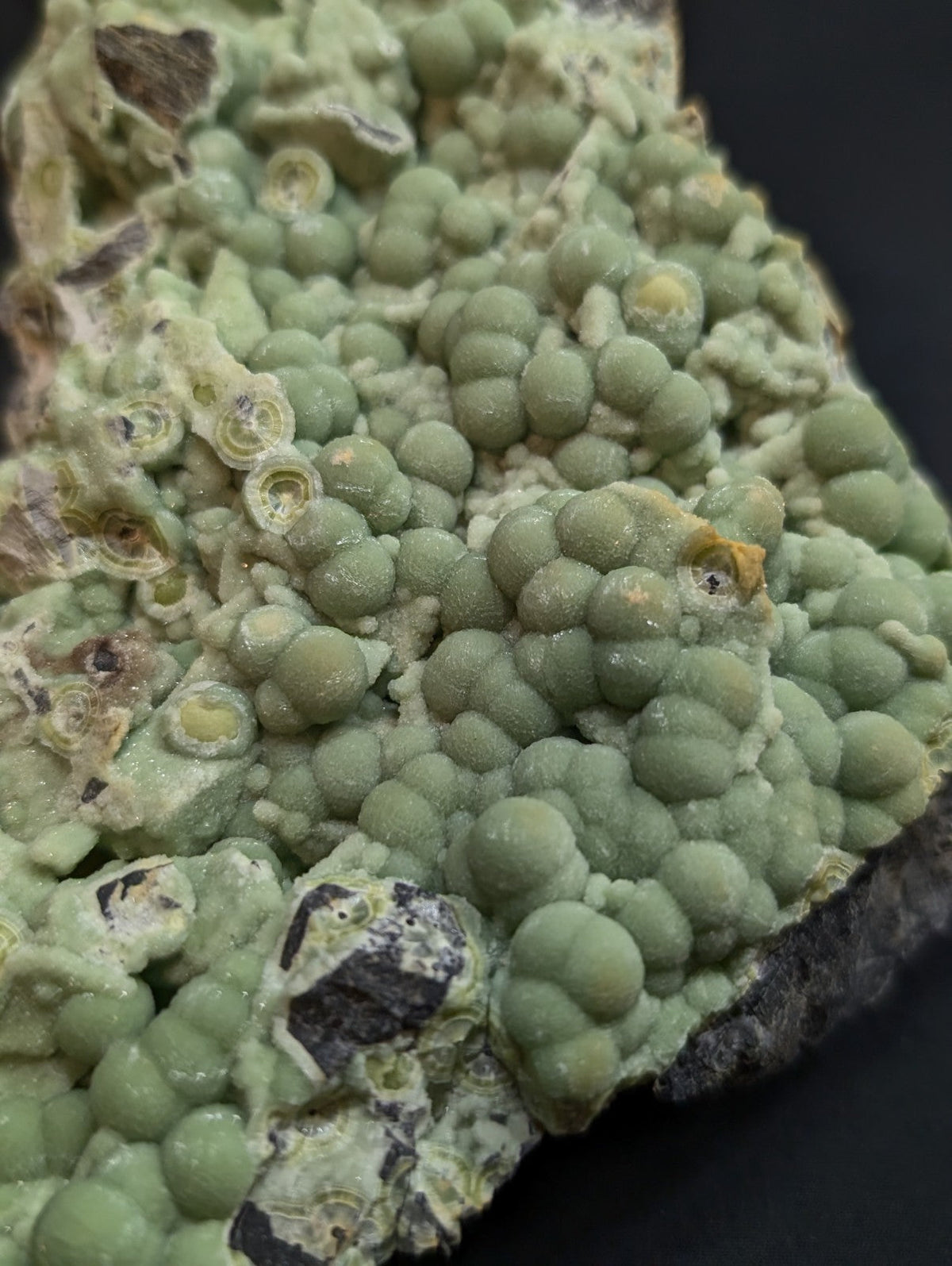 A cluster of light green, spherical mineral formations with some yellowish centers and dark patches on a rough surface.