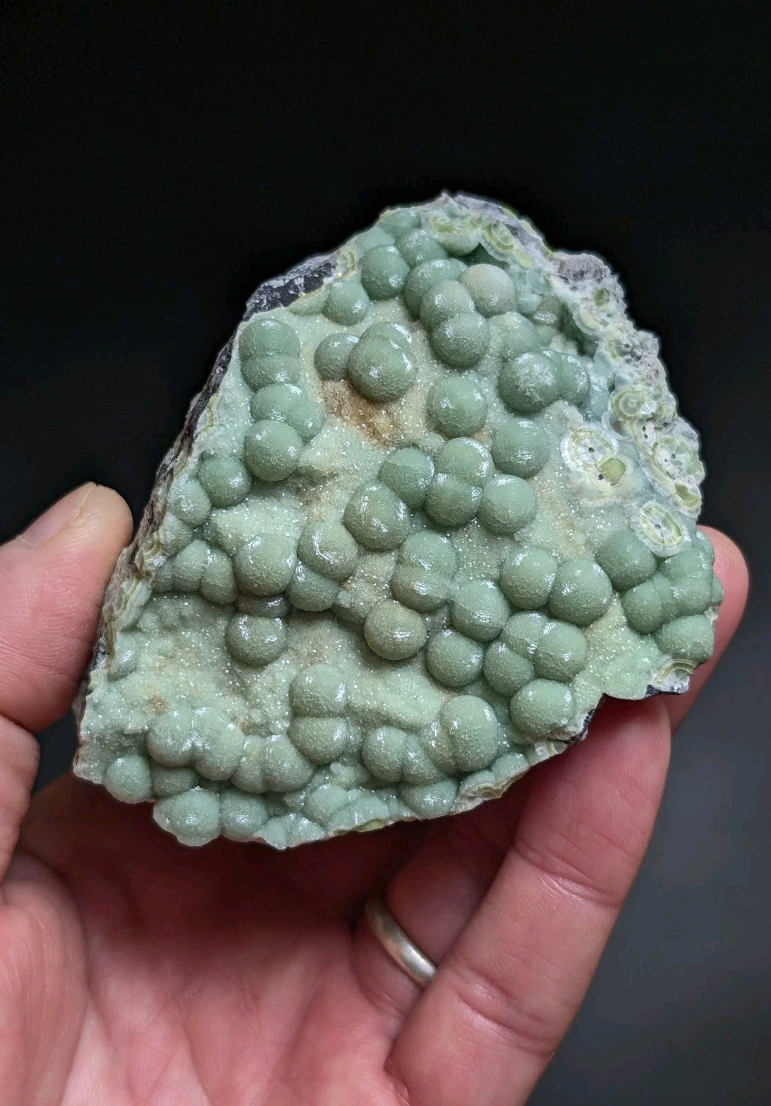 A hand holds a light green, textured mineral specimen featuring numerous rounded, bead-like formations clustered together.