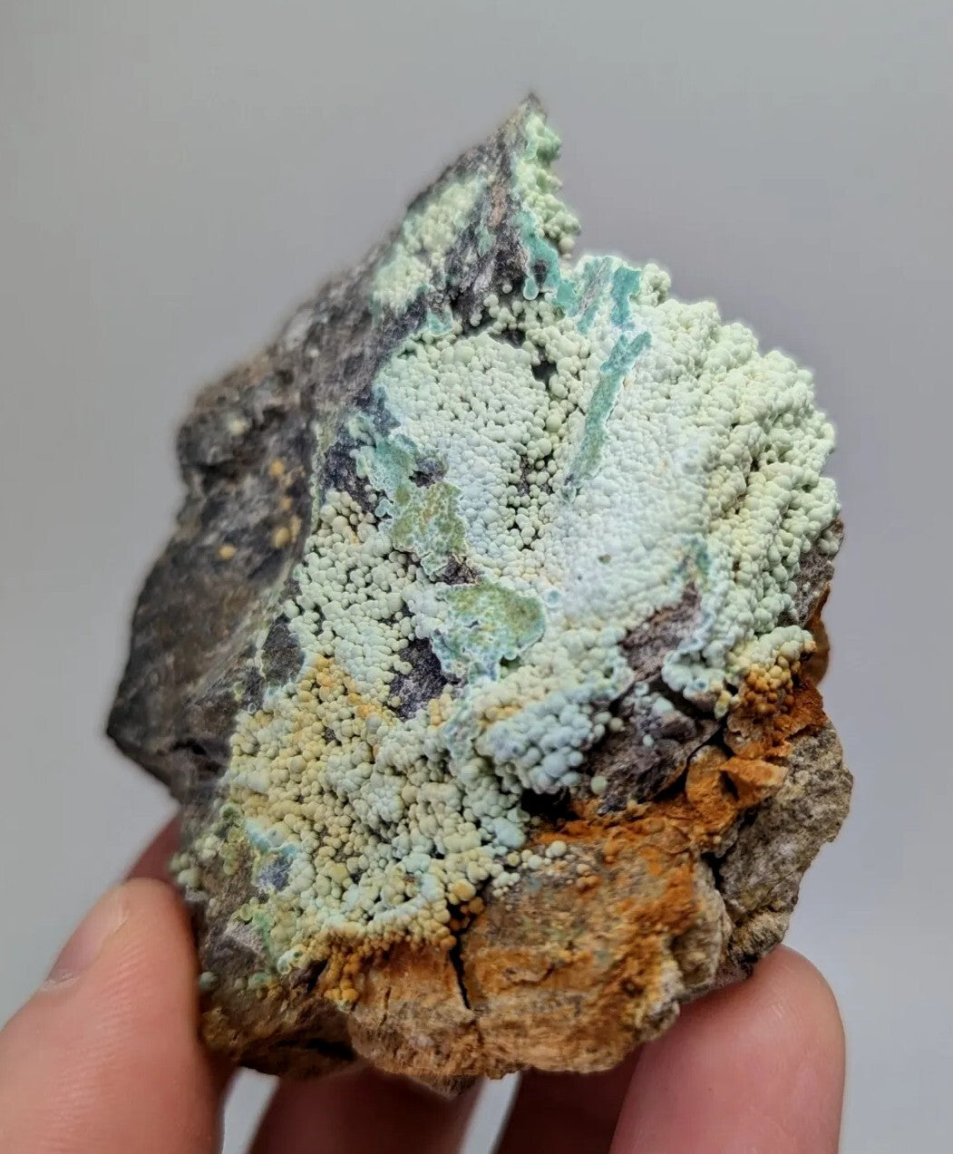 A hand holds a rugged, irregularly shaped rock specimen featuring vibrant green and pale blue mineral formations over dark gray and orange-brown base material.