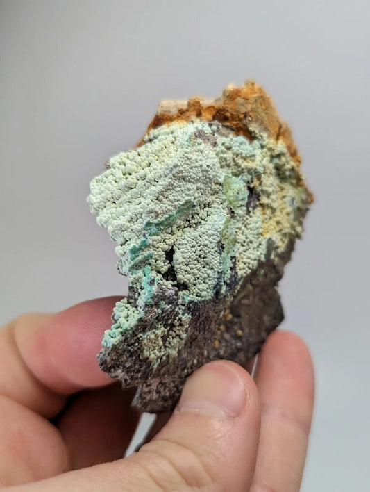 A hand holds a rough, irregularly shaped mineral specimen featuring vibrant green and pale turquoise crystalline formations over a dark brown base.